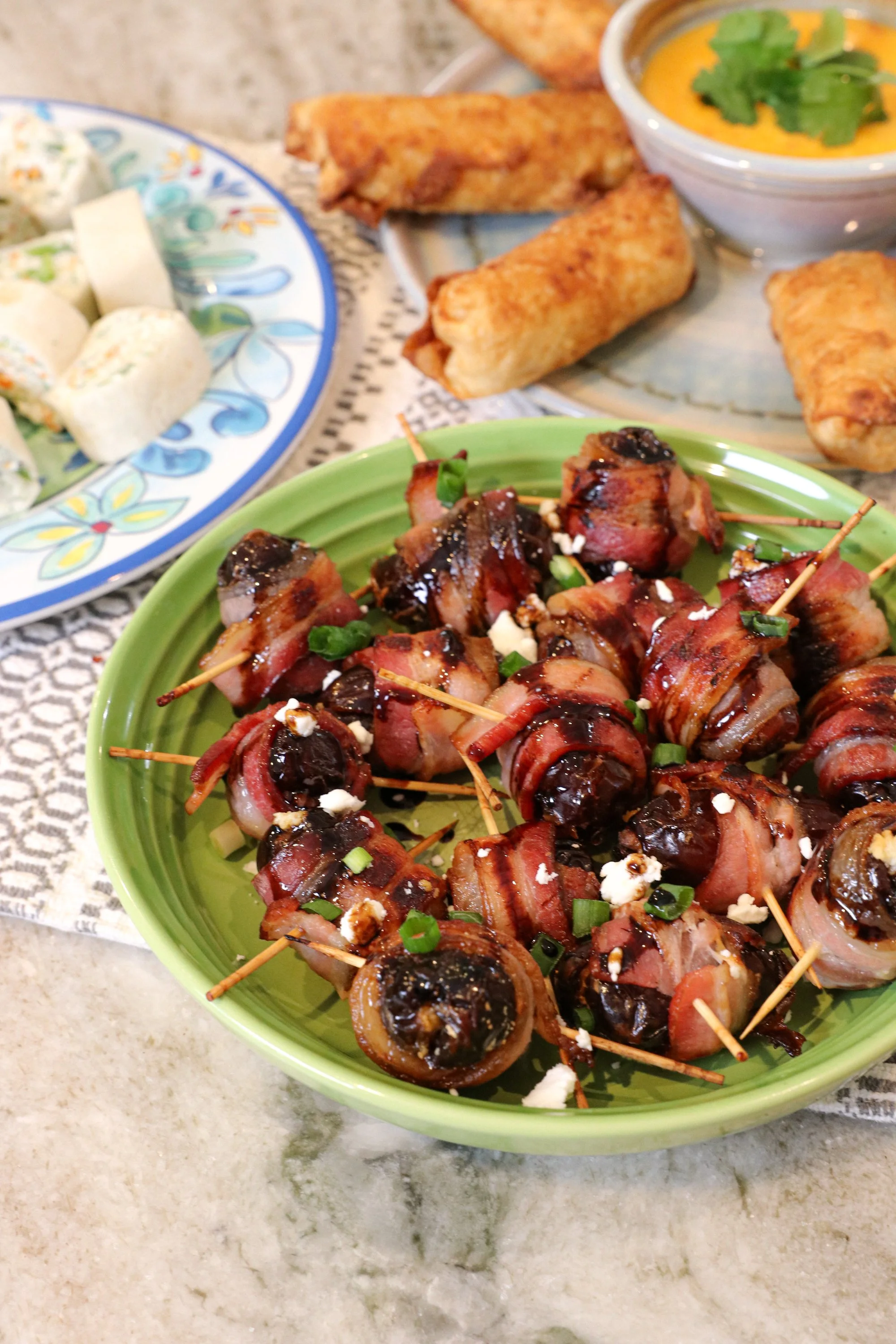 Bacon-wrapped dates topped with crumbled cheese and chopped green onions on a green plate, with spring rolls and a dipping sauce in the background.
