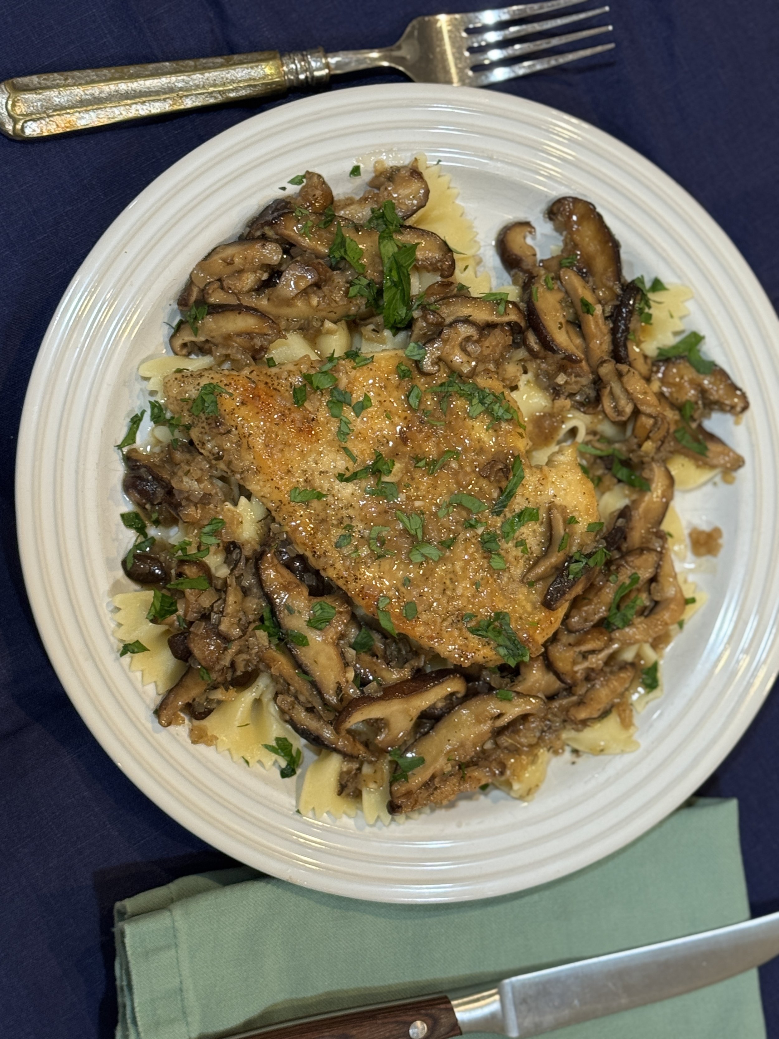 Plate of cooked bowtie pasta with sautéed mushrooms, topped with a seasoned piece of fish garnished with chopped parsley, on a decorative plate. Silver fork and wooden-handled knife rest on dark tablecloth.