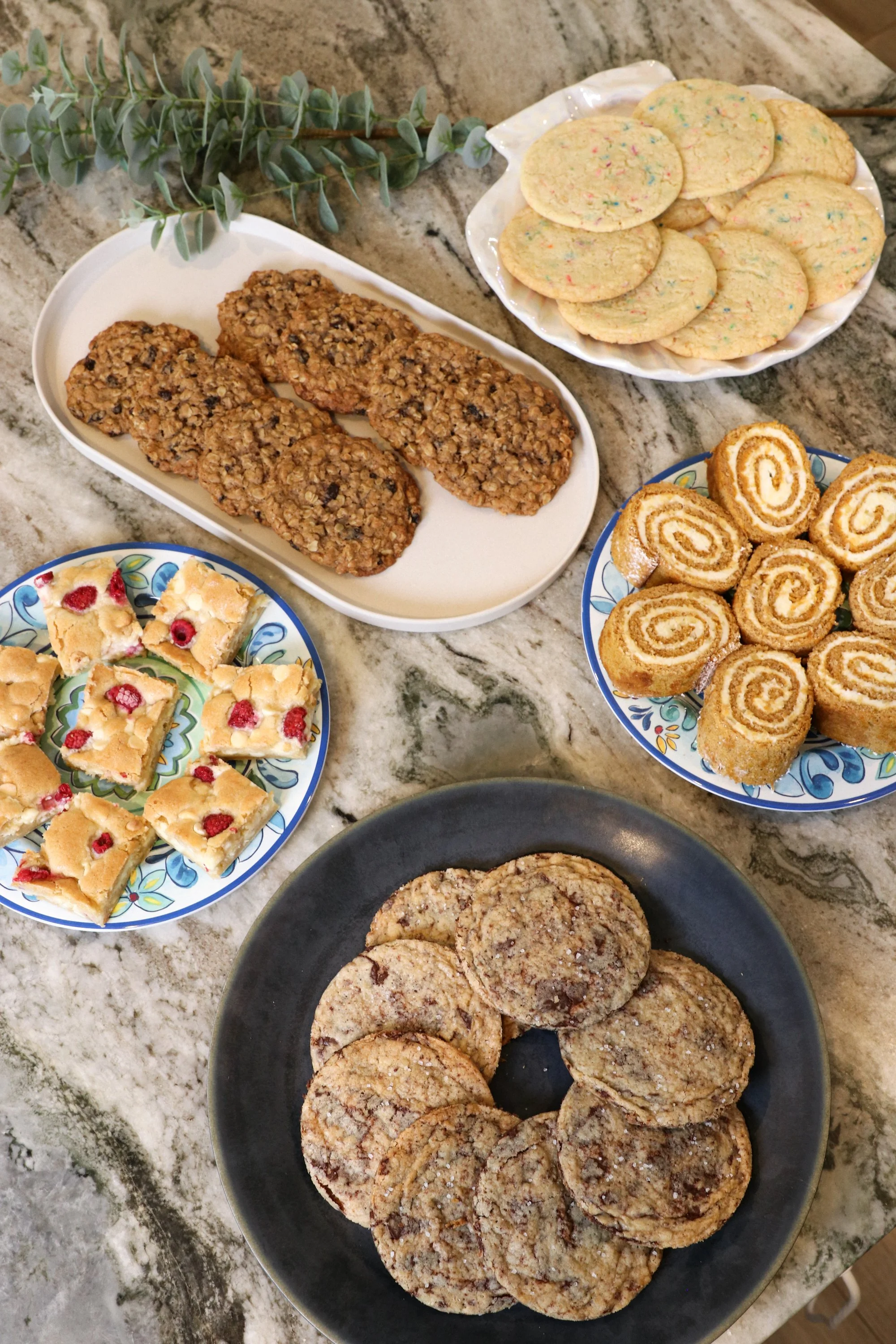 An assortment of cookies on various plates on a marble countertop, including oatmeal cookies, sugar cookies with sprinkles, rolled Swiss roll cookies, raspberry bars, and chocolate chip cookies.