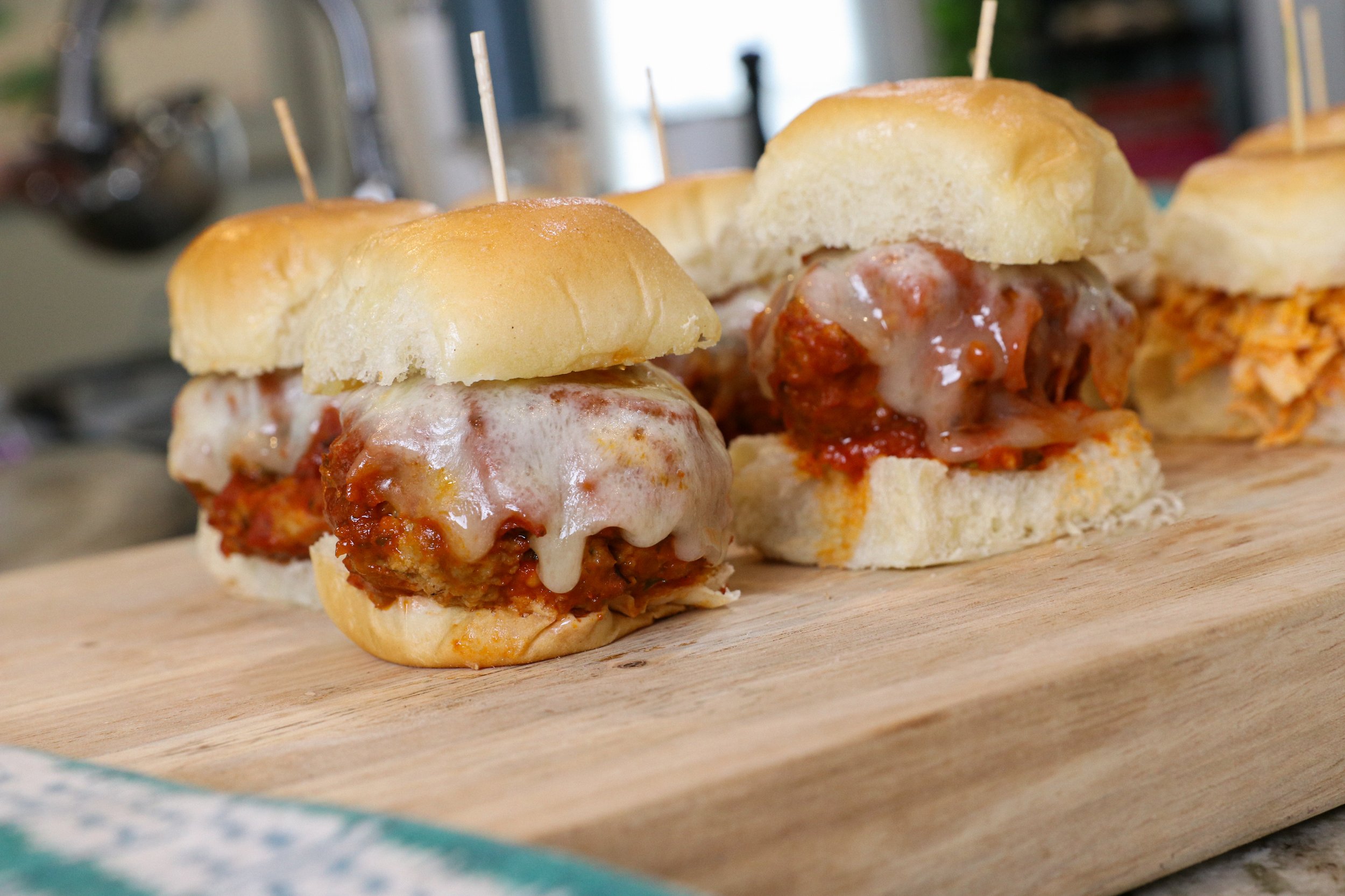 Three mini meatball sandwiches with melted cheese on a wooden cutting board in a kitchen.