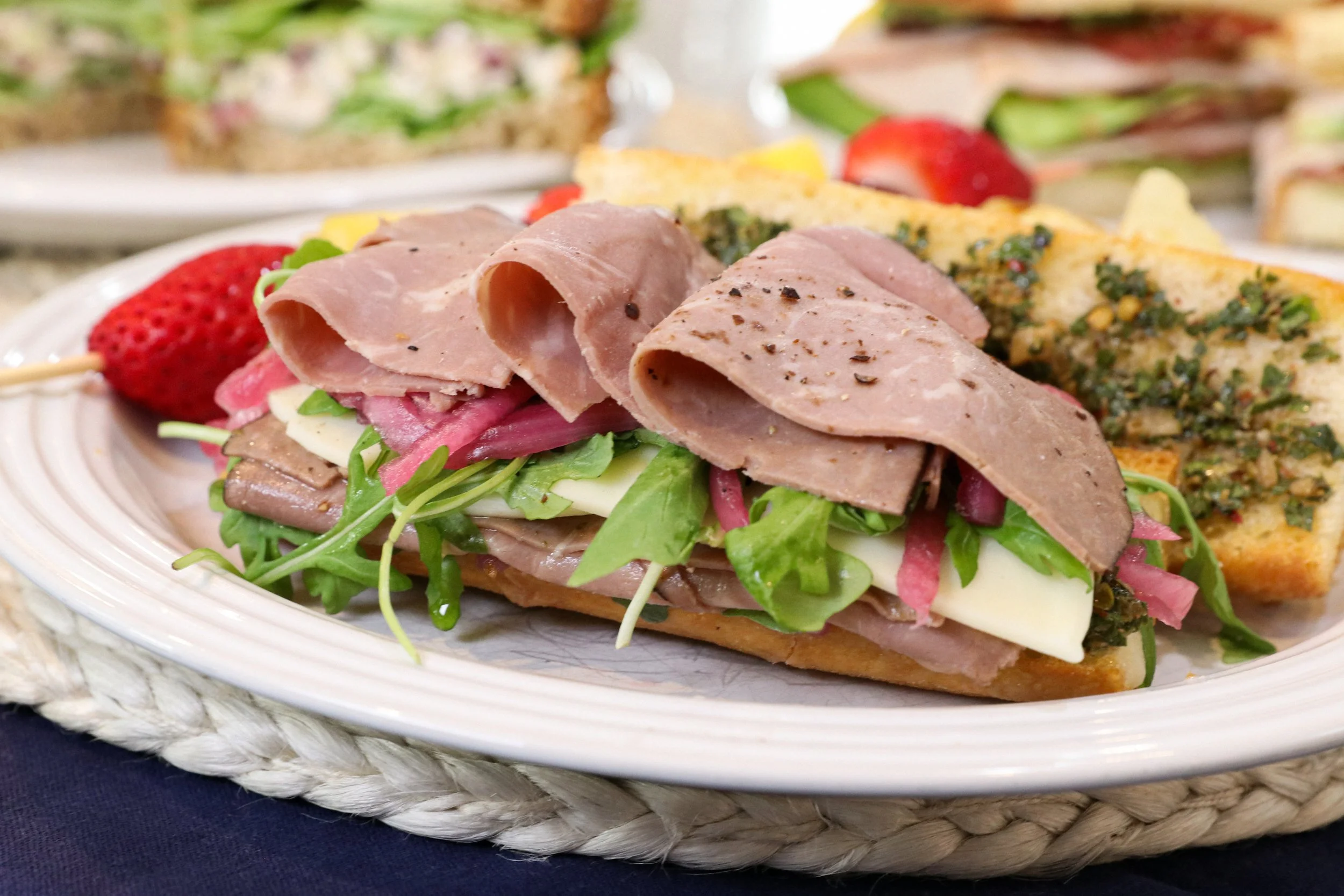 Open-faced sandwich with sliced roast beef, arugula, cheese, pickled onions, and radishes on bread, with a strawberry and a slice of garlic bread with herbs in the background.