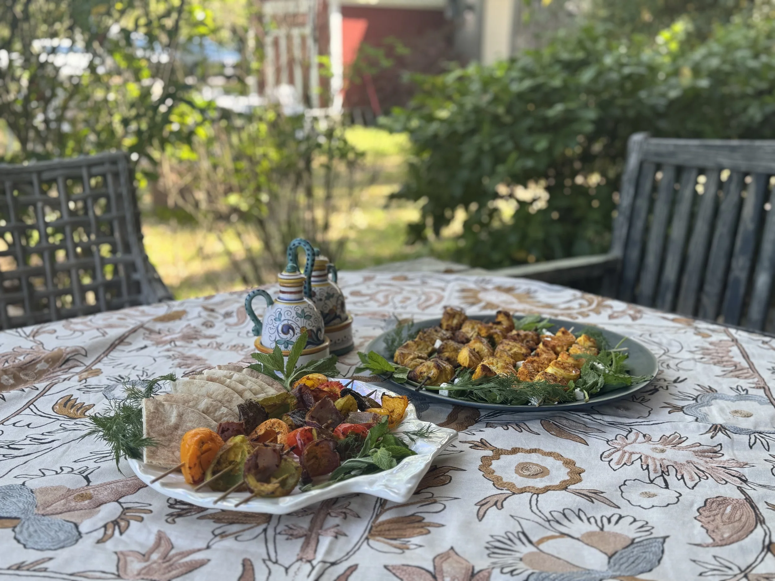 Outdoor table set with plates of grilled vegetables and meat skewers, surrounded by greenery with additional chairs in the background.