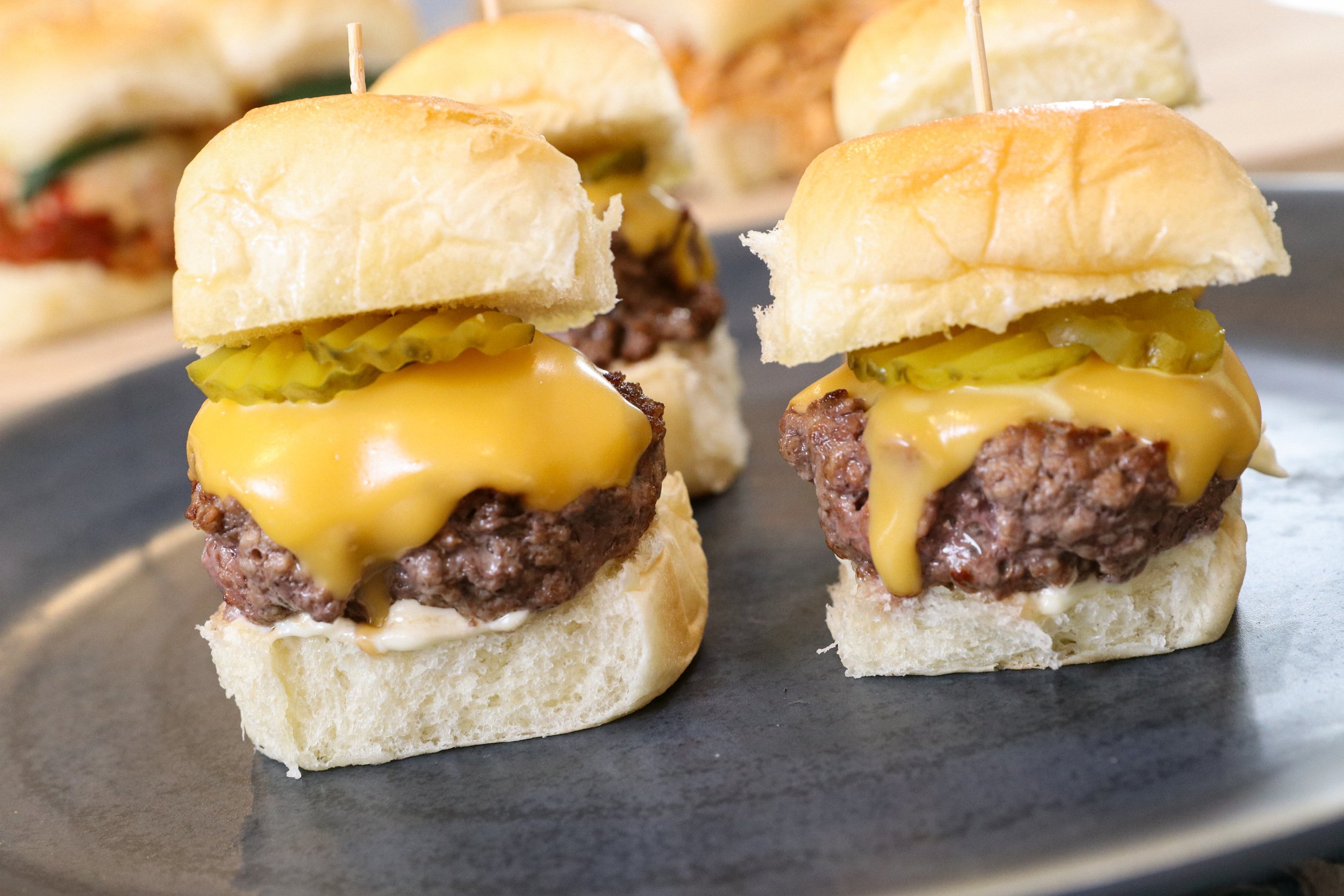 Mini cheeseburgers with pickles on a dark platter.