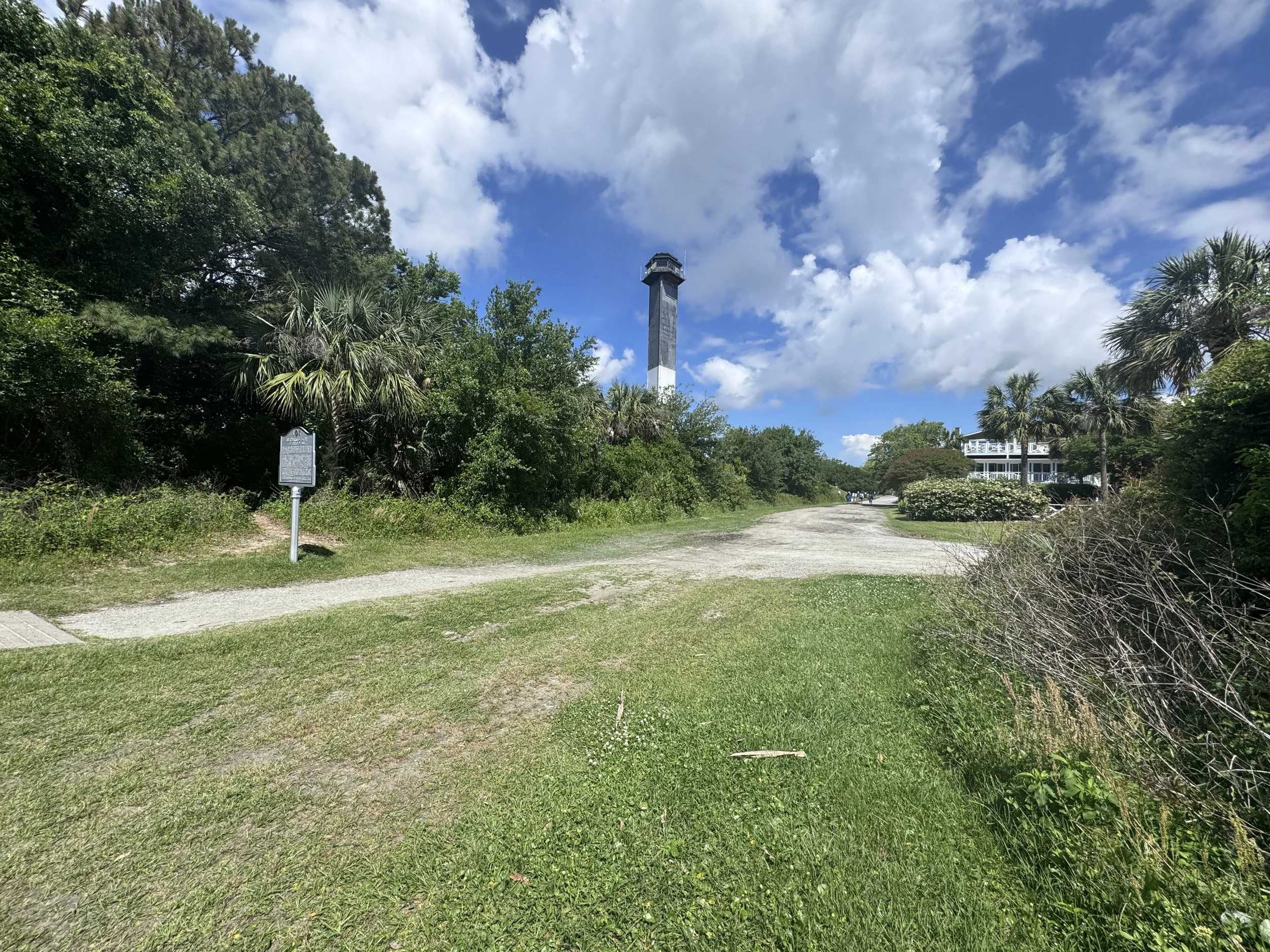 A gravel path leading towards a tall black and white striped lighthouse, surrounded by greenery and blue sky with white clouds.