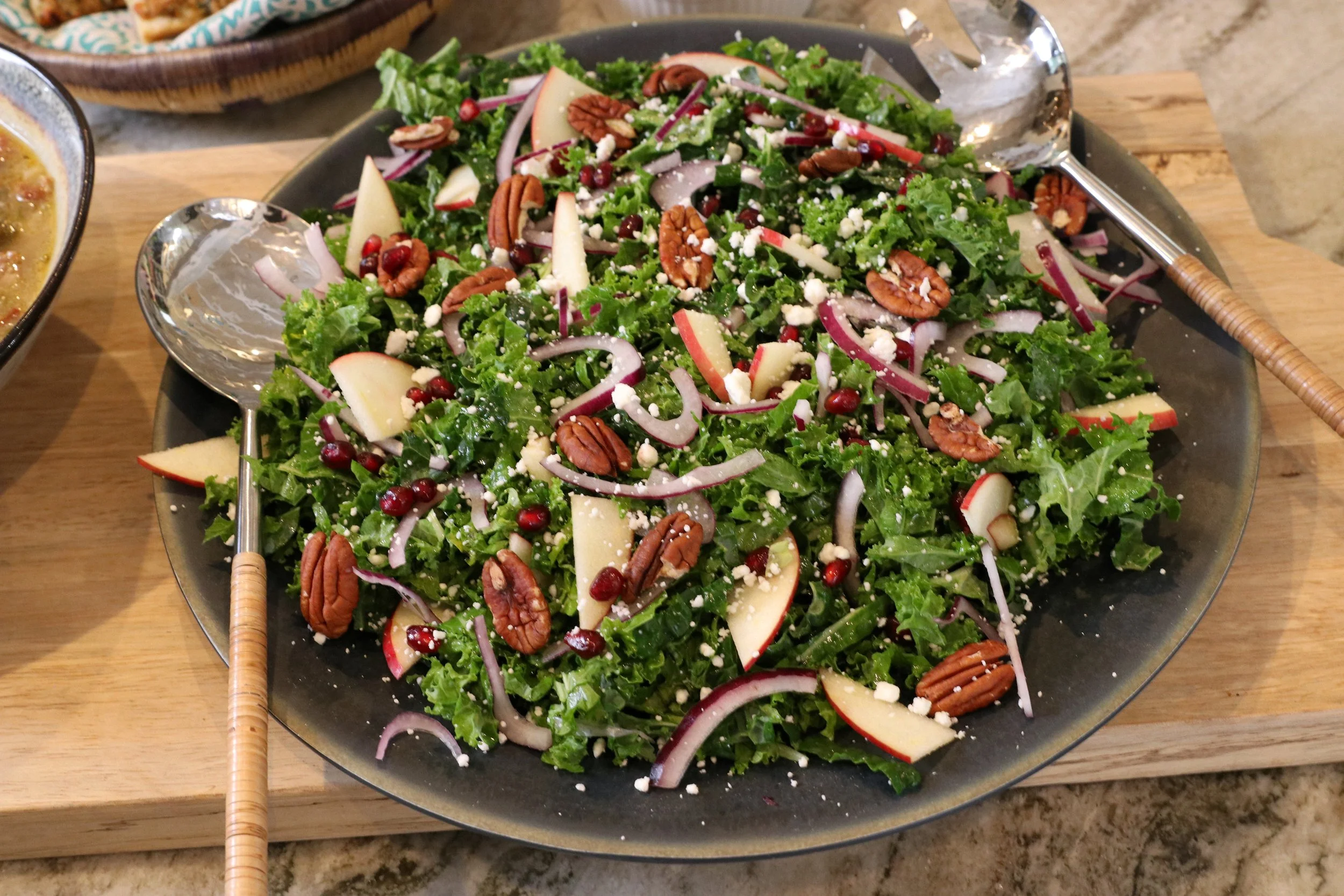 A plate of fresh mixed greens salad with sliced apples, red onions, pecans, pomegranate seeds, and crumbled cheese, served with tongs on a wooden board.