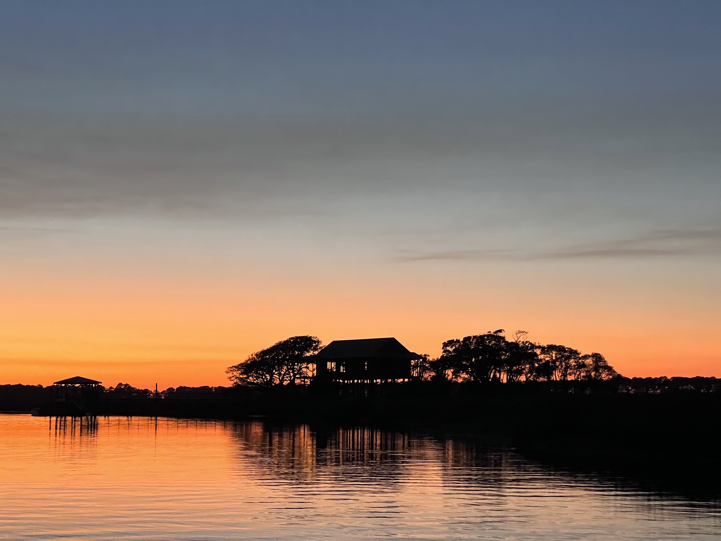 Silhouette of house and trees along a waterway at sunset with gradient sky from orange to blue and calm reflections on the water.