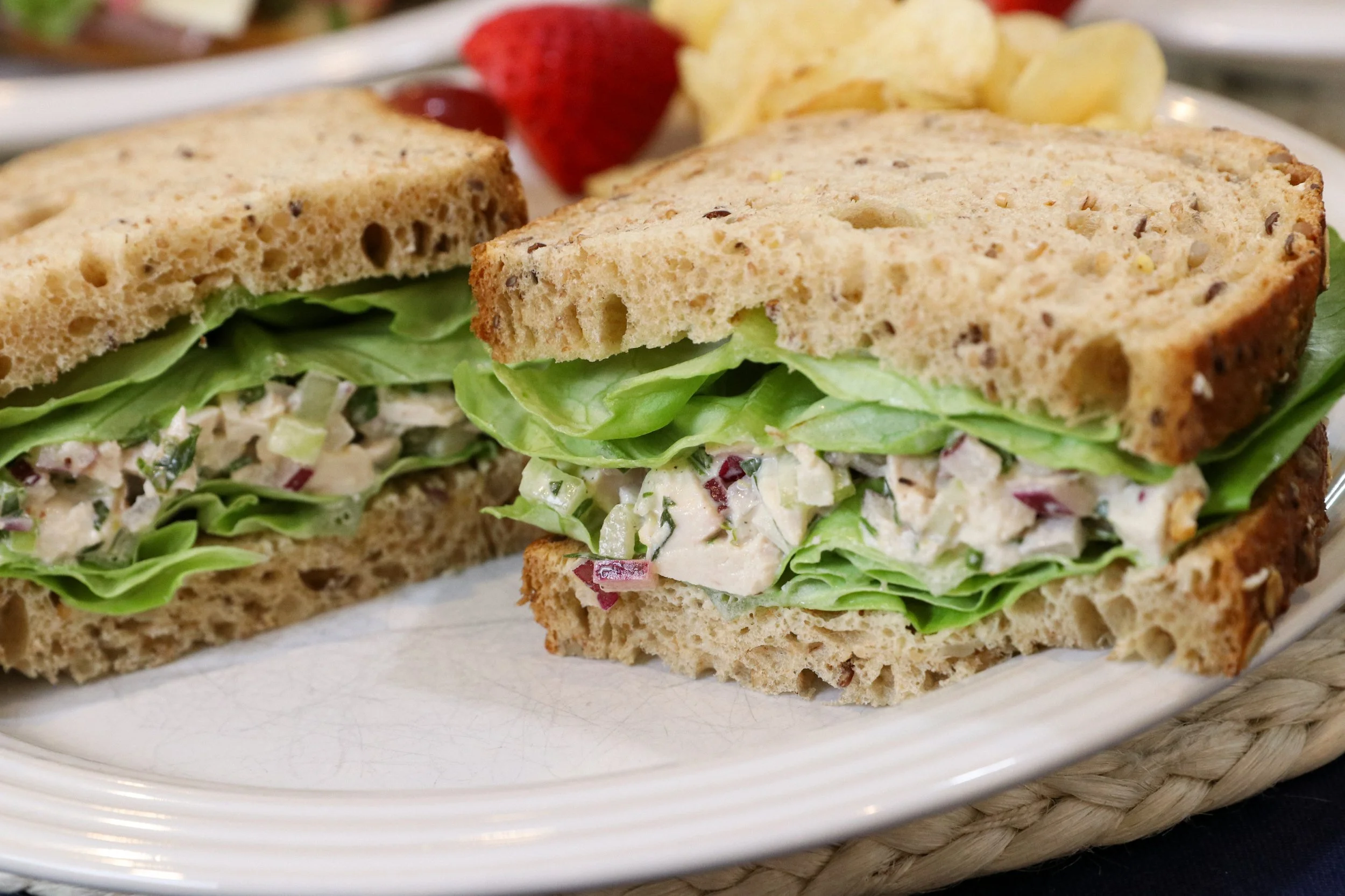 Close-up of a chicken salad sandwich on multigrain bread with lettuce, served on a white plate with potato chips and strawberries in the background.