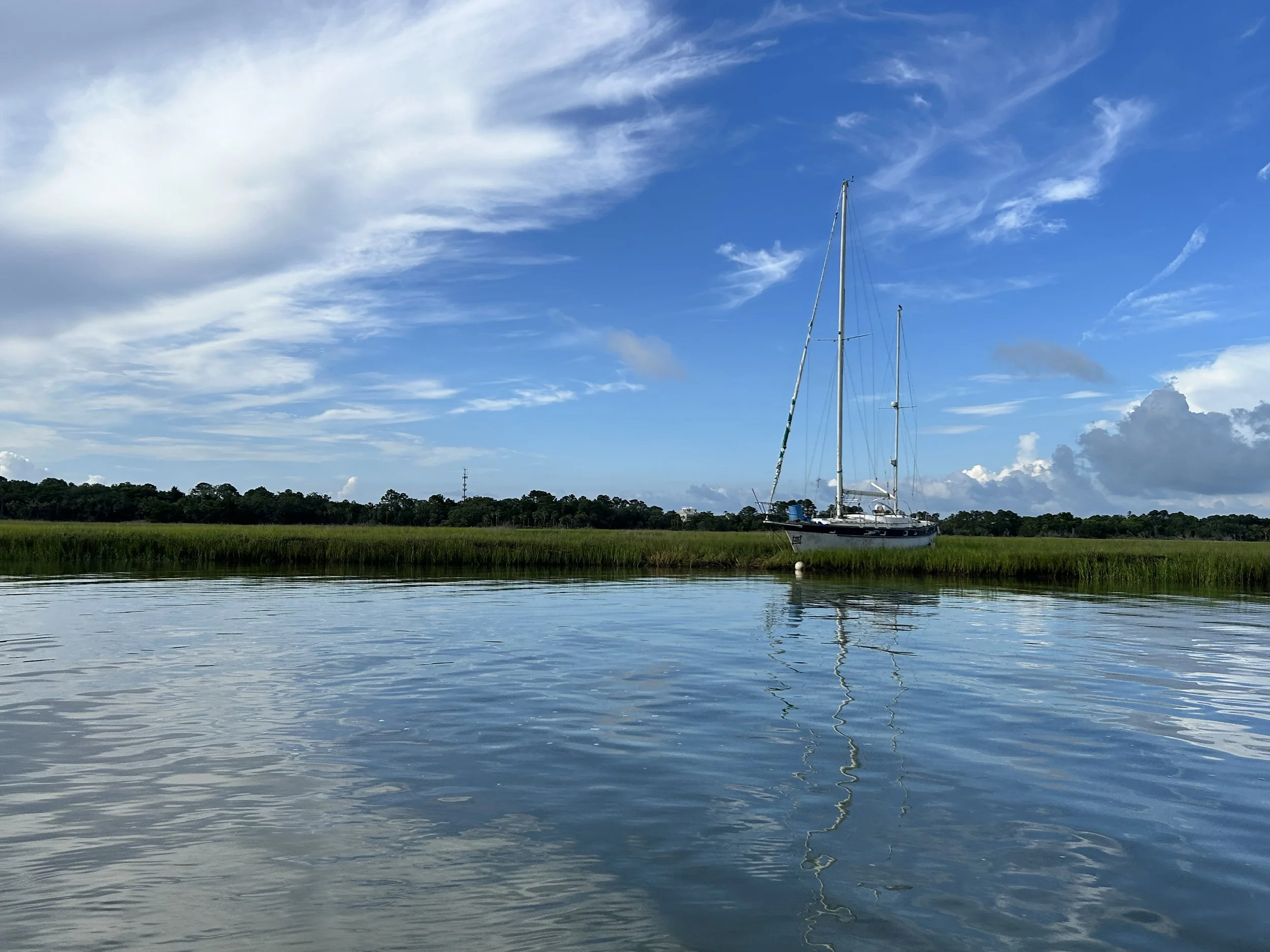 Calm water with a grassy shoreline in the background, a sailboat anchored near the shore, under a partly cloudy blue sky.