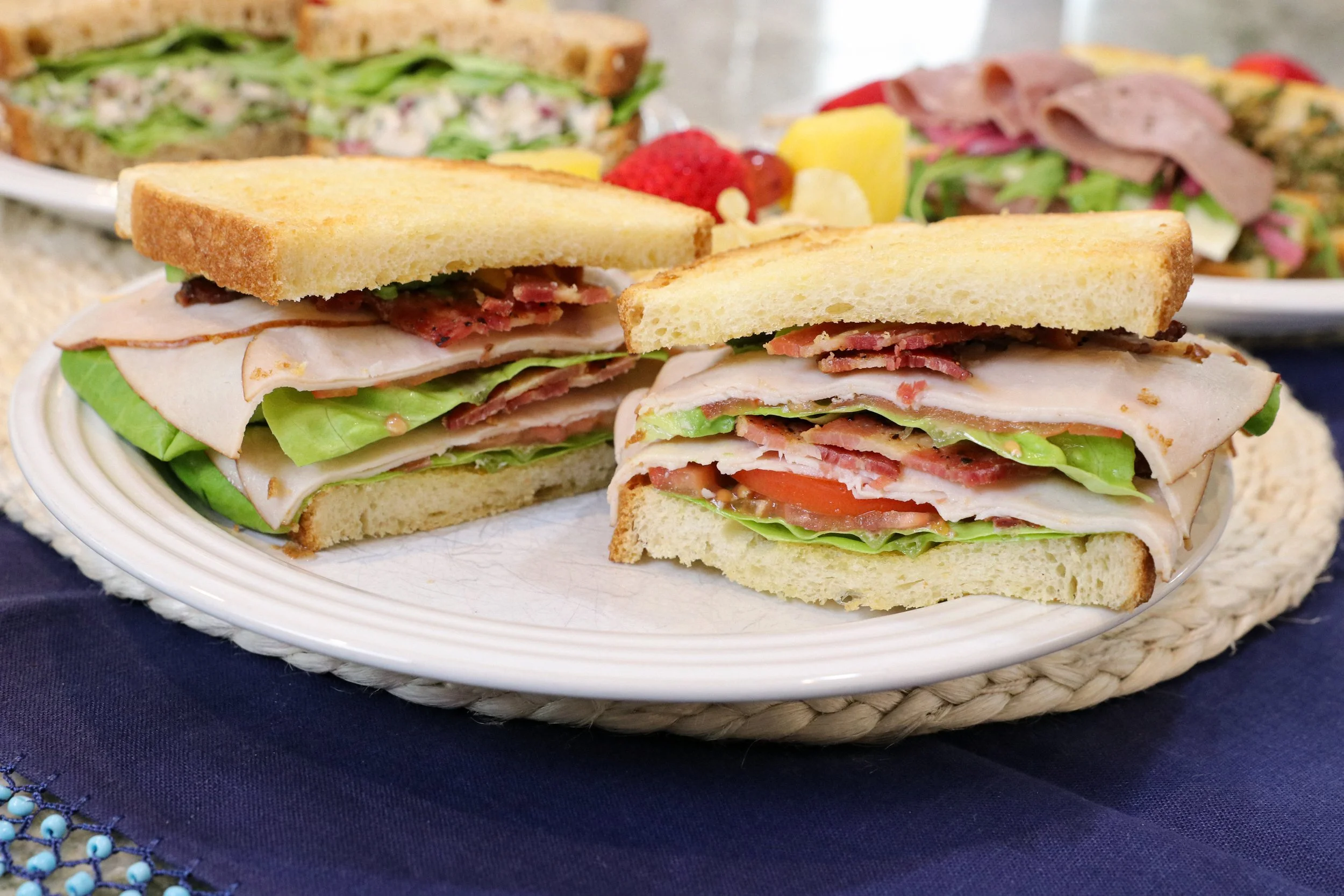Close-up of a plate with two sandwich halves filled with turkey, lettuce, tomato, bacon, and slices of bread, on a blue tablecloth with other plates of sandwiches in the background.