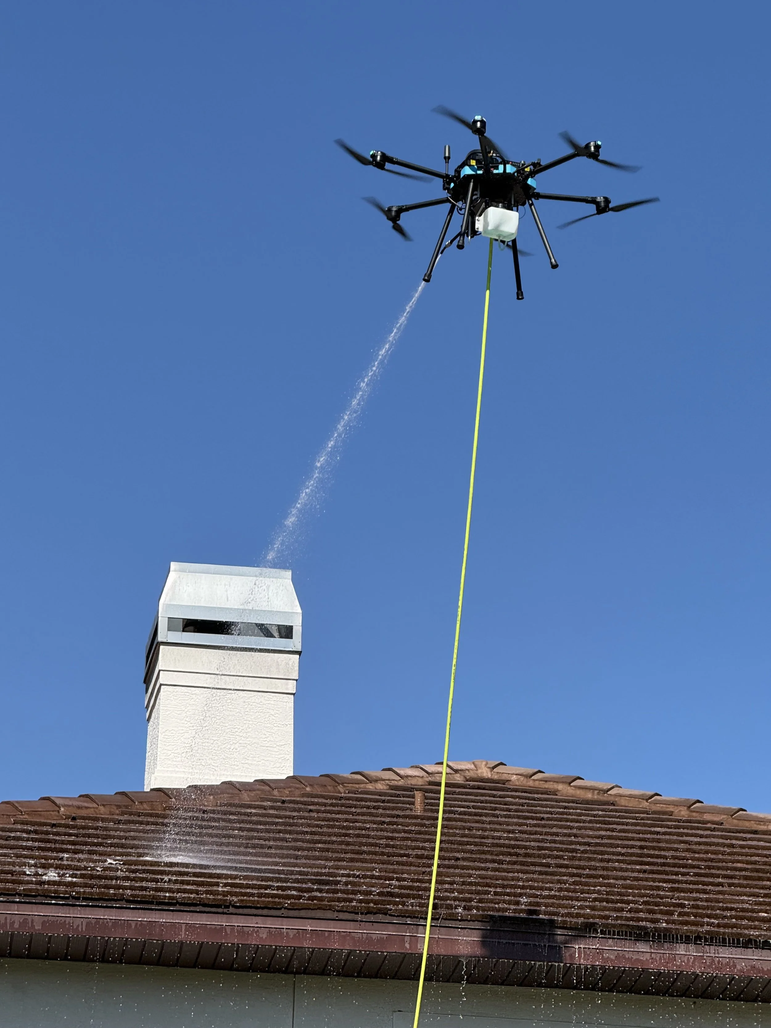 A drone with multiple propellers is flying above a house's roof, spraying water or a liquid substance near a chimney on a sunny day with clear blue sky.