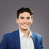 Young man smiling, wearing a blue blazer and white shirt, against a gray background.