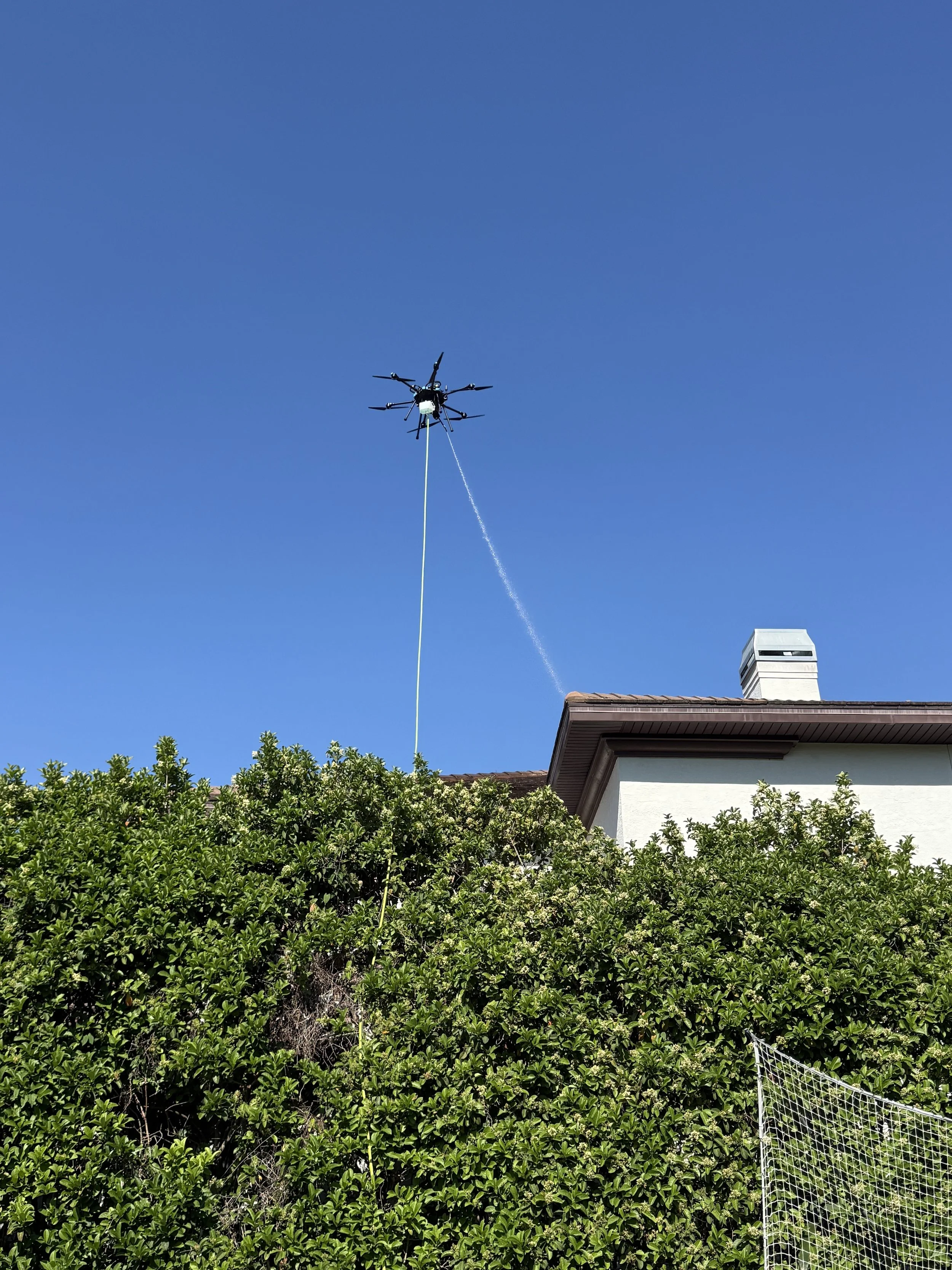 A drone flying above a house with a long tether, in front of green bushes and a clear blue sky.