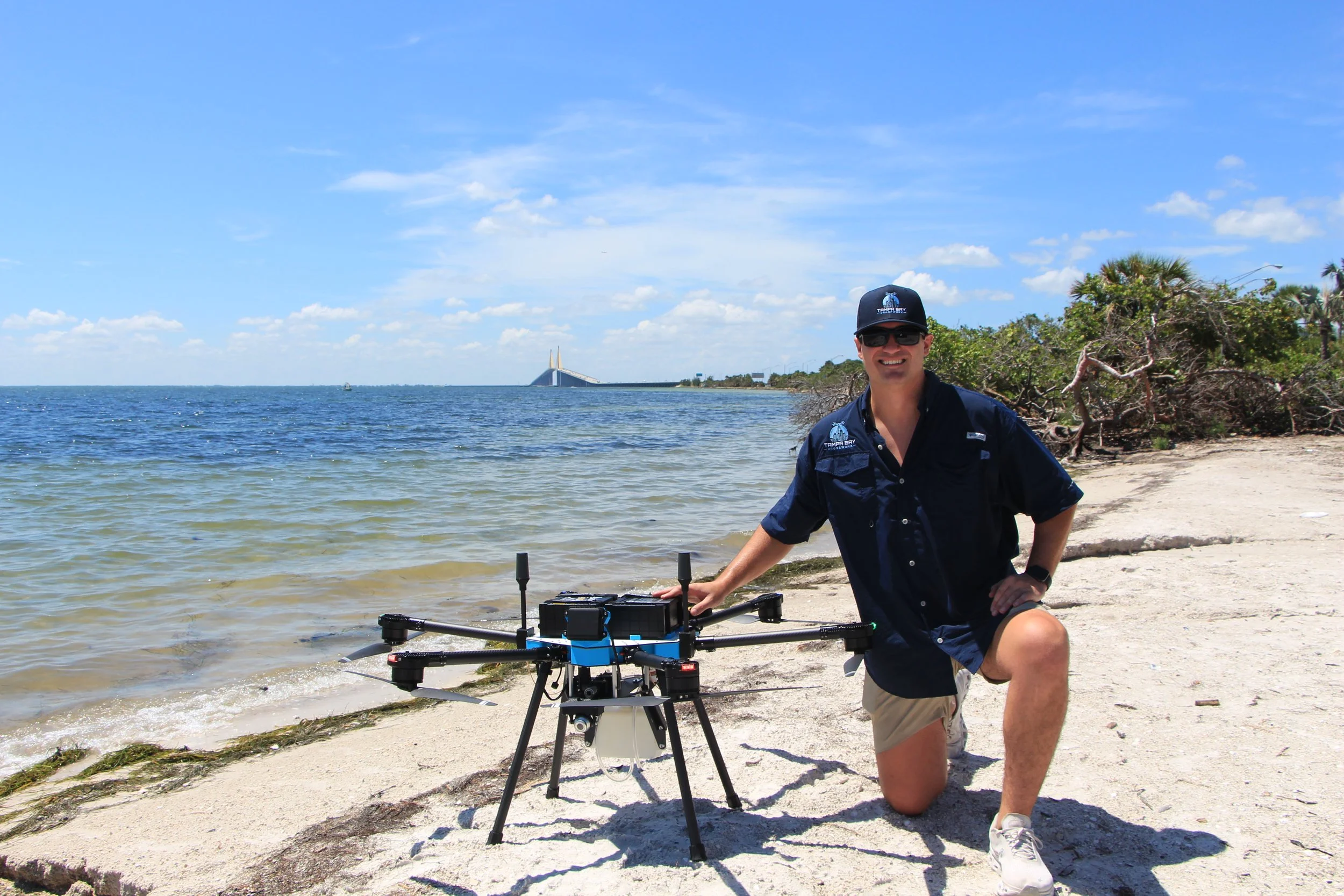 A man kneeling on a sandy beach with water and shoreline behind him. He is wearing sunglasses, a navy shirt, and khaki shorts, and is smiling at the camera. There is equipment on a stand in front of him, and the background includes a blue sky, some trees, and a bridge in the distance.
