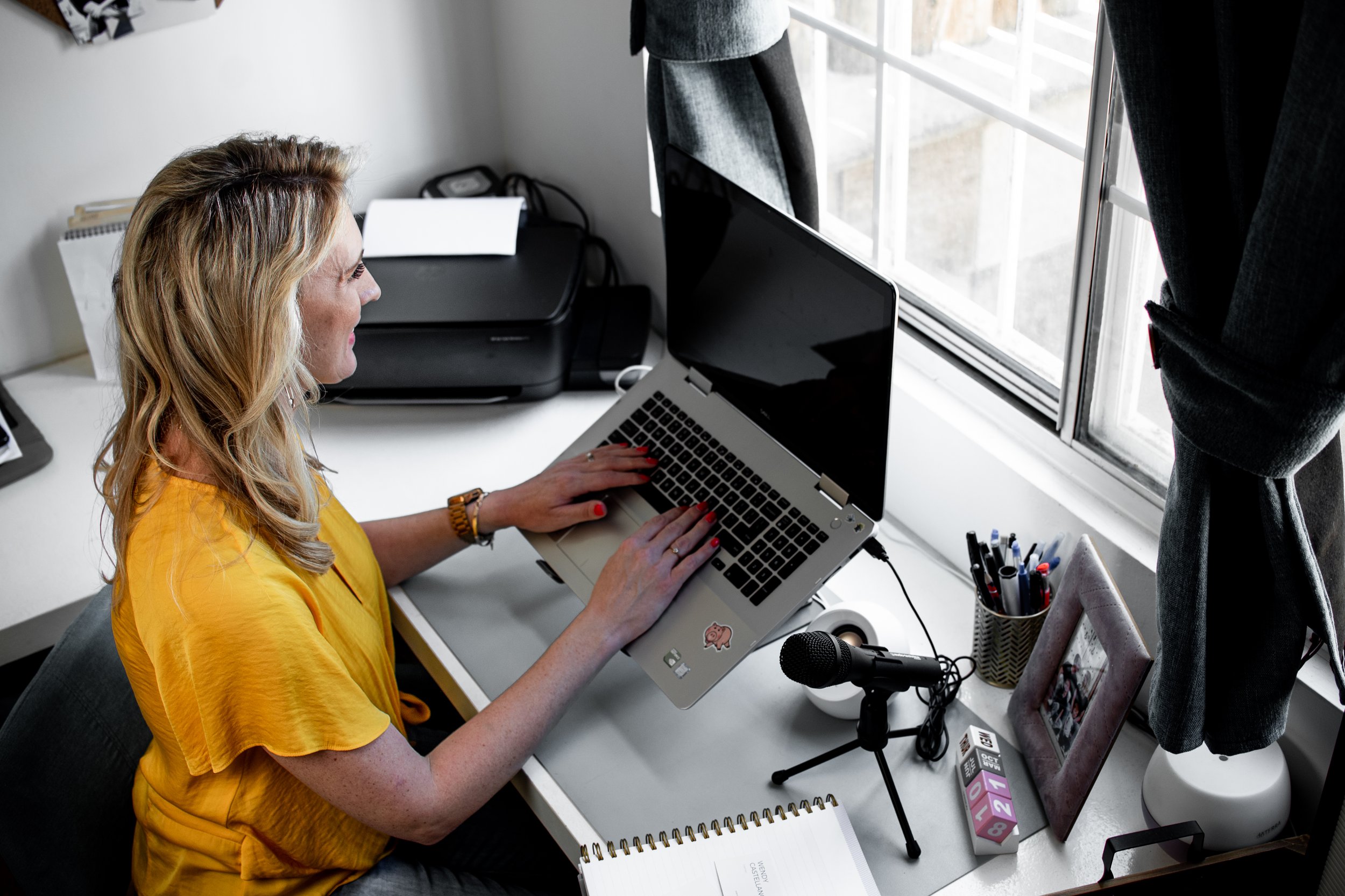 A woman with blonde hair and a yellow top sitting at a desk, typing on a laptop. The desk has a microphone, a notebook, pens in a holder, a printer, and a window nearby with gray curtains.