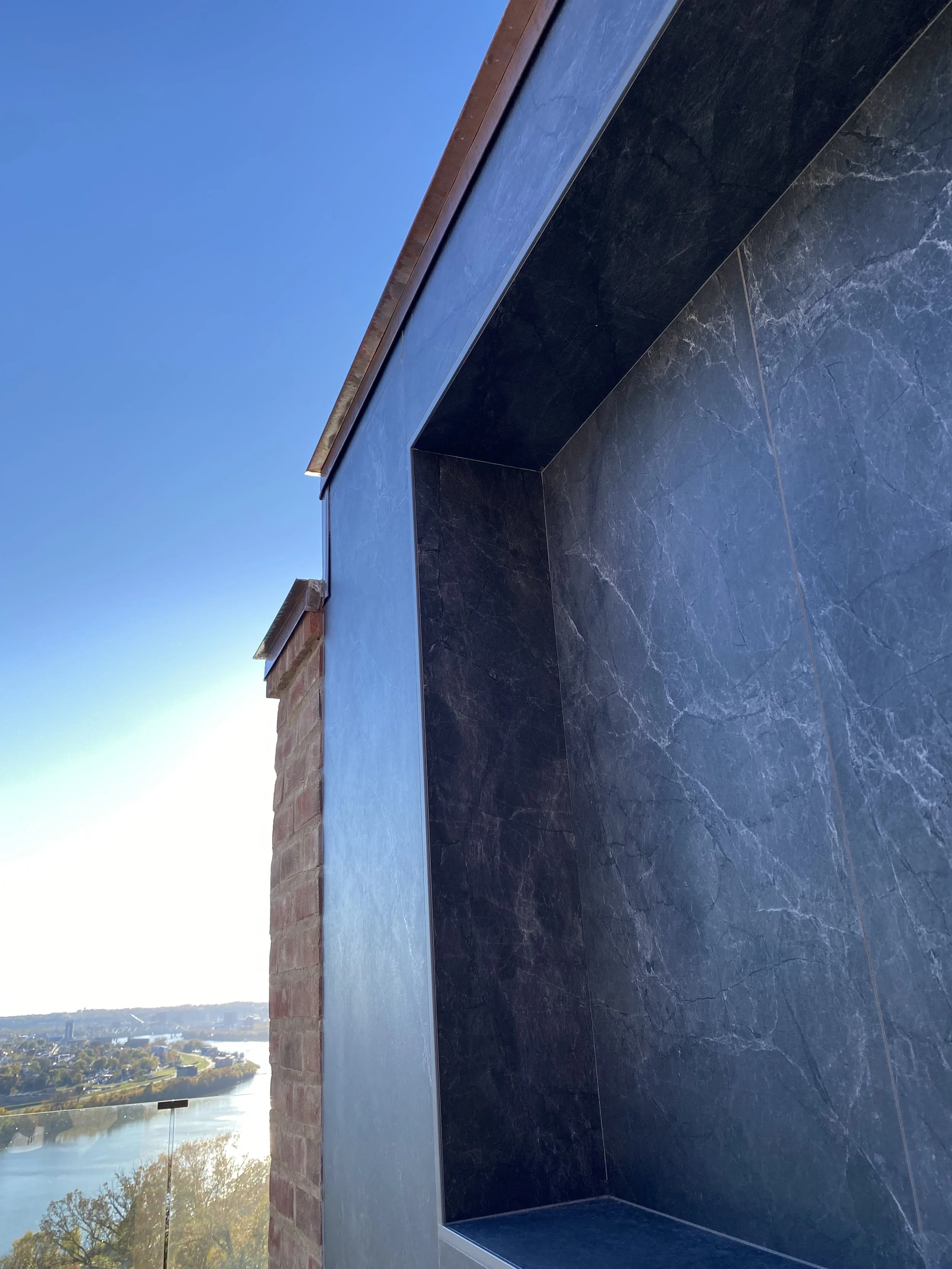 Close-up view of a dark marble or stone wall with a glass balcony overlooking a river and cityscape under clear blue sky.