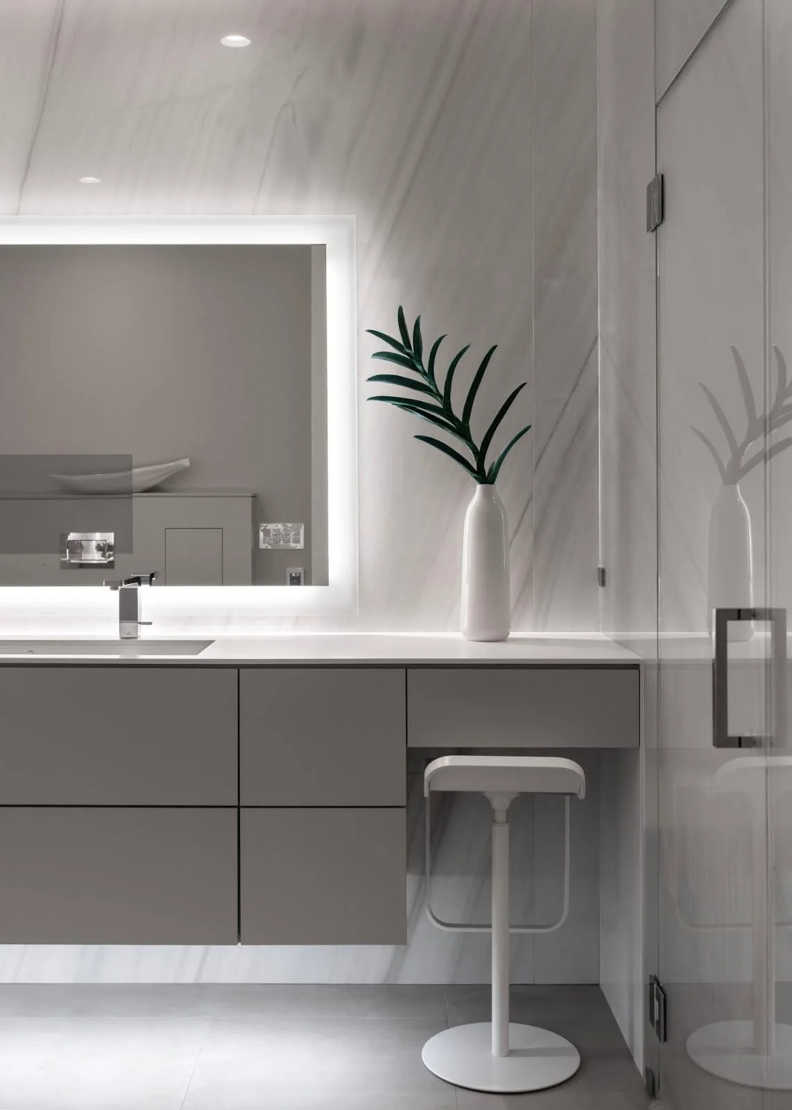 Modern bathroom with grey cabinetry, illuminated mirror, white countertop, and a tall white vase with a green plant, all reflected in a mirror and glass shower door.