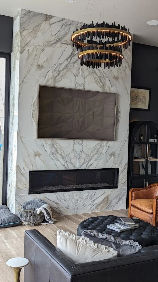 Modern living room with a marble feature wall, a television, a fireplace, and a black leather couch. A distinctive circular black and gold chandelier hangs from the ceiling.