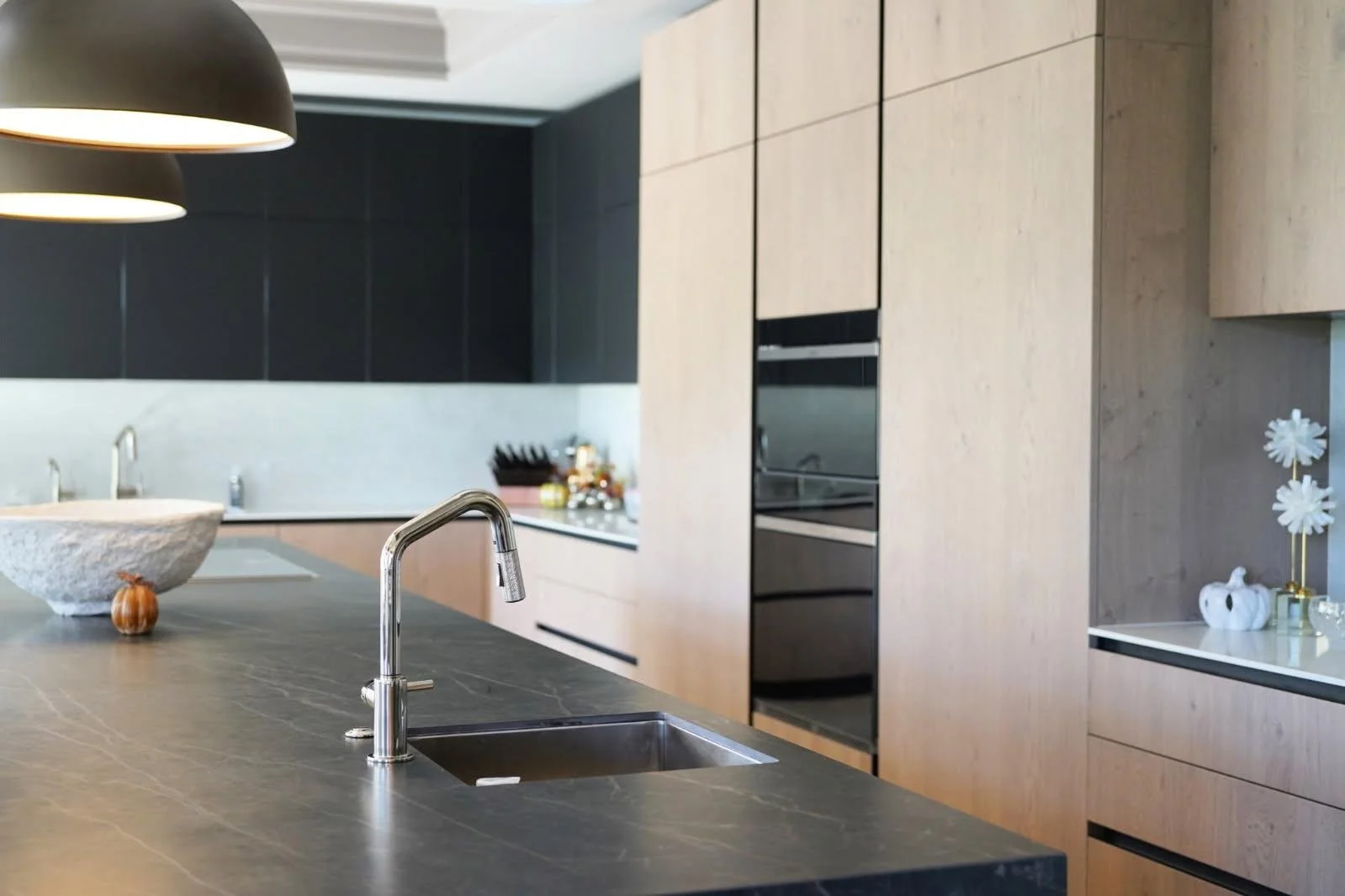 Modern kitchen with black marble countertop, a built-in sink, and wooden cabinets. Pendant lights hang above the island, and there are decorative items on the right side of the image.