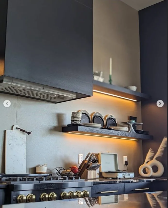 Modern kitchen with black cabinets, grey backsplash, and open shelving displaying dishes and decorative items, illuminated by warm under-shelf lighting.