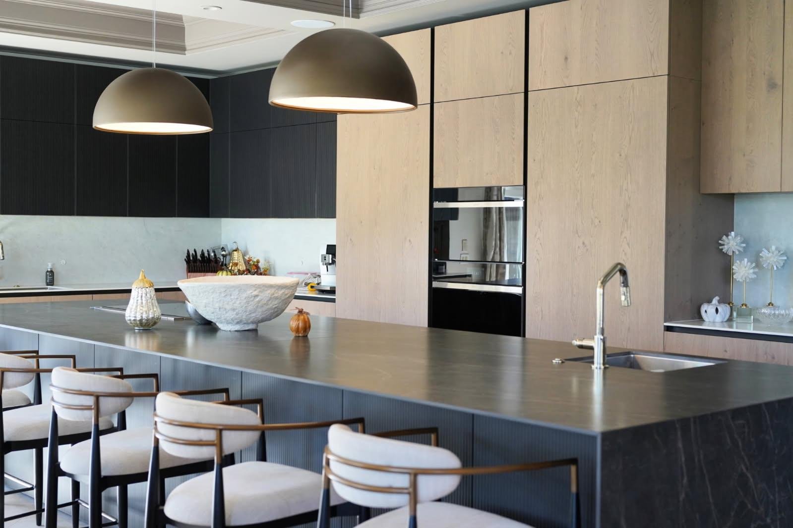Modern kitchen with a large gray island, black and wood cabinets, and two pendant lights. There are chairs around the island, decorative items on the counter, and artwork on the wall.