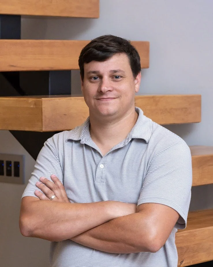A man with short dark hair, wearing a light gray and white striped polo shirt, standing with arms crossed in front of a wooden staircase.