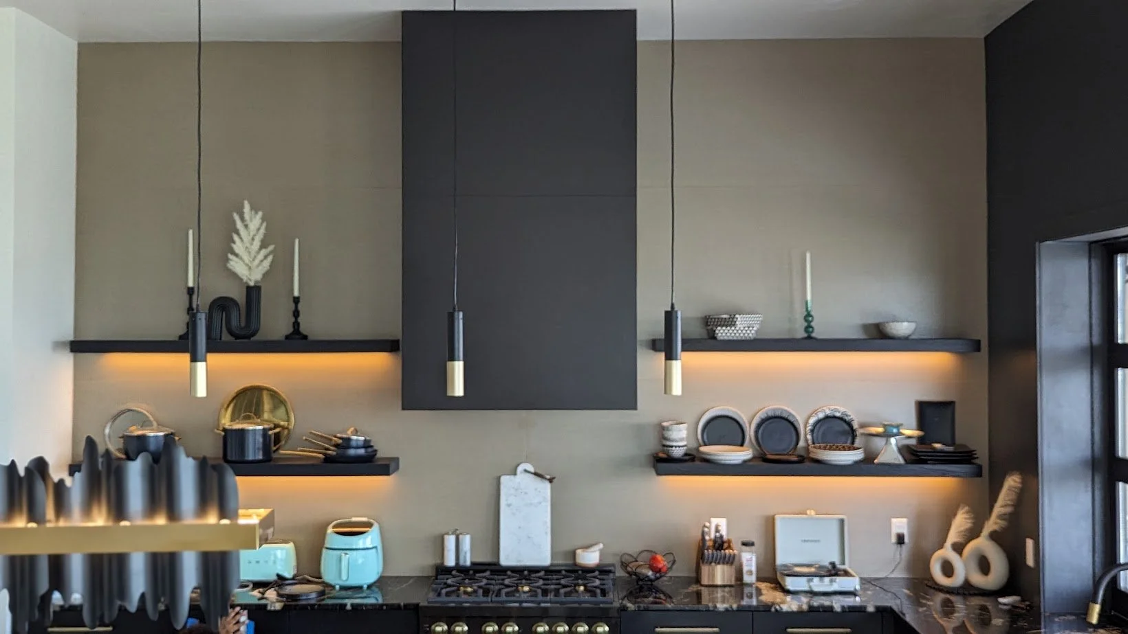 Modern kitchen with black shelves and a black stove, decorated with dishes, bowls, and kitchen appliances. Two black pendant lights hang over the counter. There are shelves with decorative items and a black cabinet in the center.