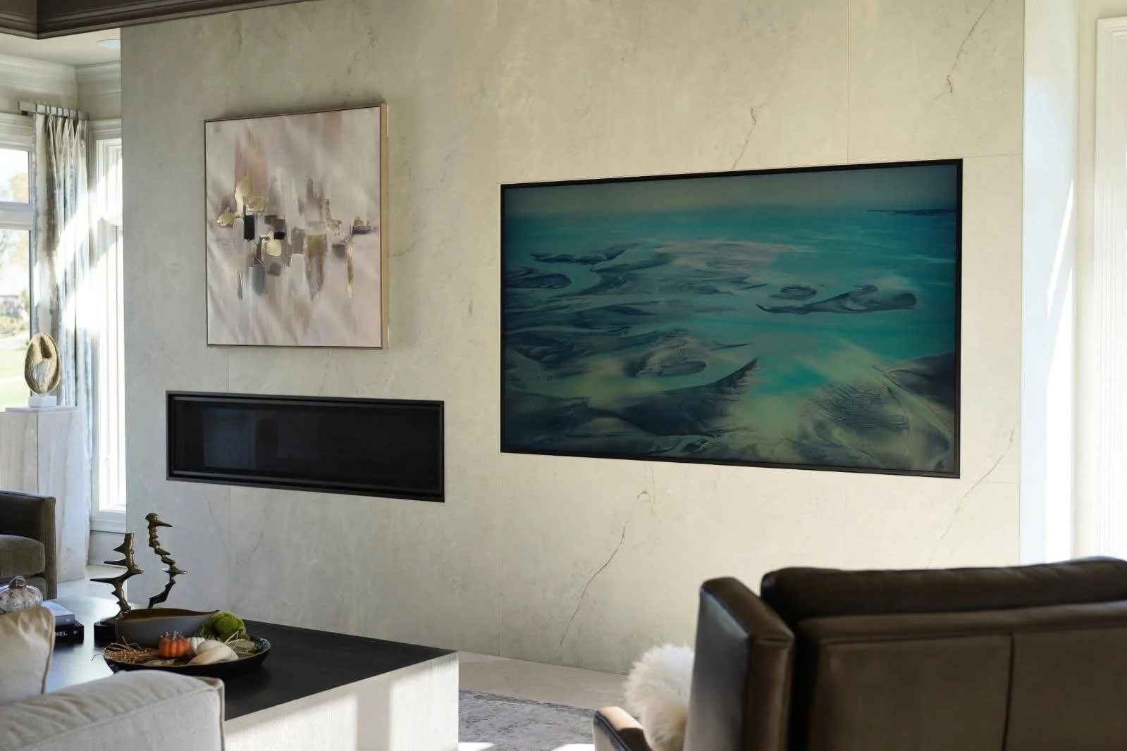 Living room with a beige wall decorated with two abstract paintings, a large flat-screen TV displaying a nature scene, and modern furniture including a beige sofa, a brown armchair, and a black coffee table with decorative items.