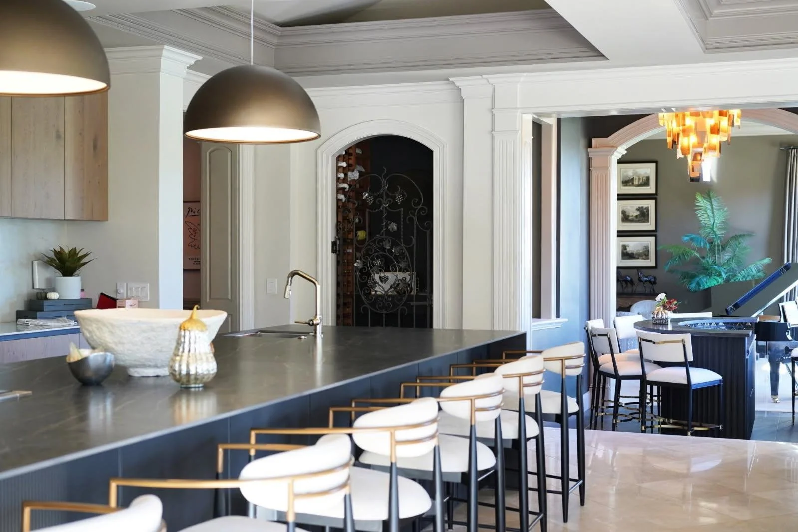 Modern kitchen with a large island, white chairs with gold accents, pendant lights, a black grand piano, and a sitting area with framed pictures and a large plant.