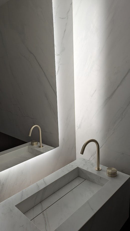 A minimalist bathroom sink with a rectangular marble basin, a sleek gold curved faucet, a small round container, and a large mirror reflecting the sink and wall.