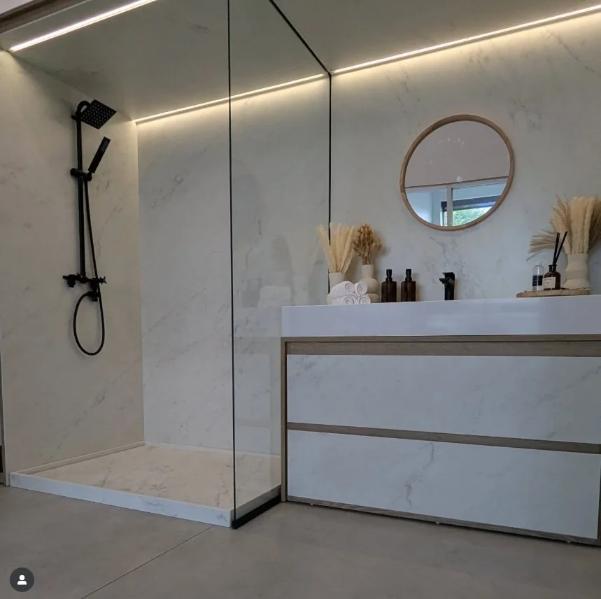 Modern bathroom with marble wall and floor, featuring a glass-enclosed shower with black fixtures, a white vanity with a round mirror, and decorative elements like dried plants and toiletries.