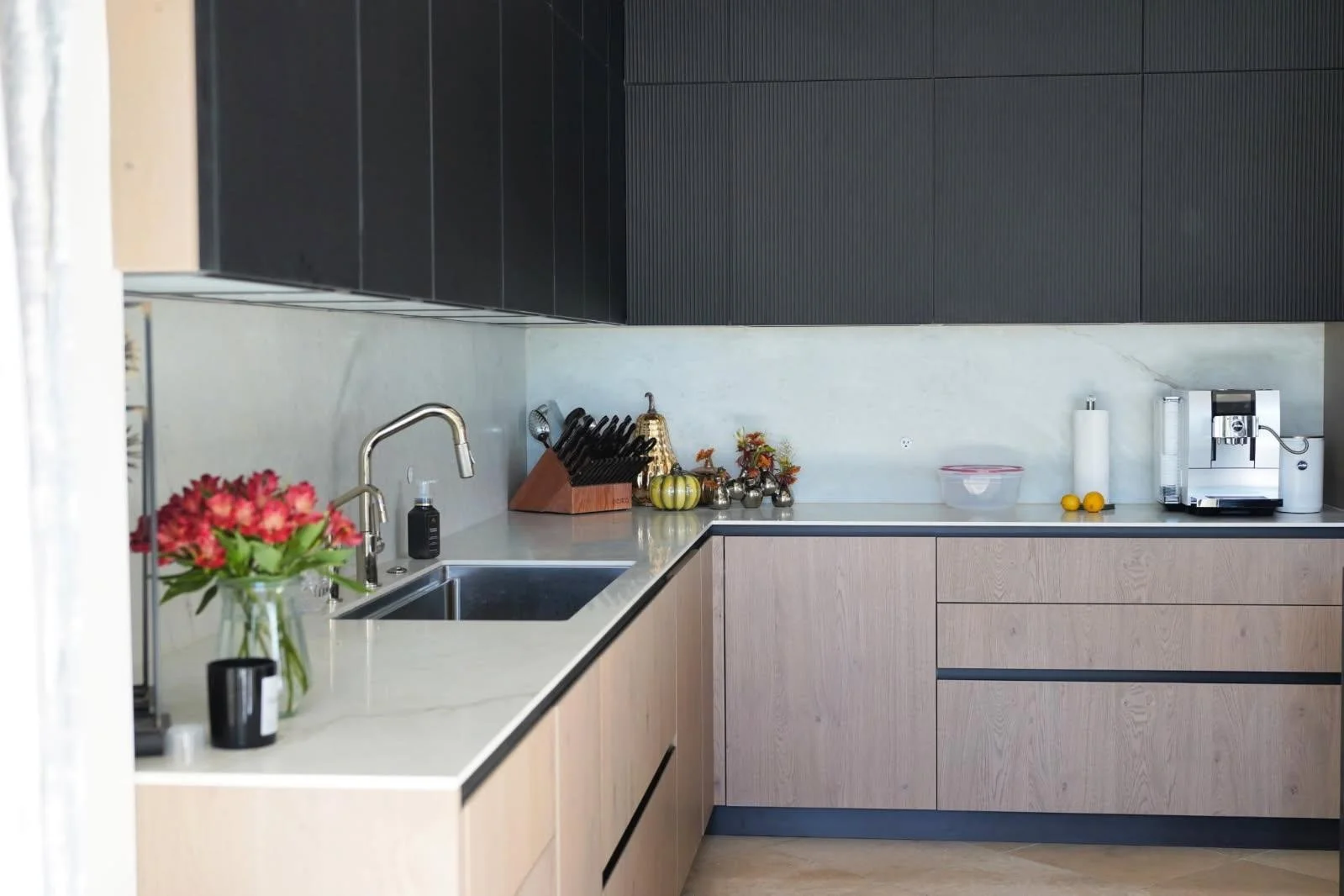 Modern kitchen with black and wooden cabinets, a marble countertop, stainless steel sink, fresh flowers, and small kitchen appliances.