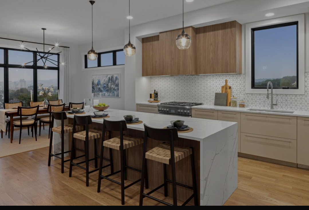 Modern kitchen with white marble island, wooden cabinets, and black chairs, adjacent to a dining area with a large window and view.