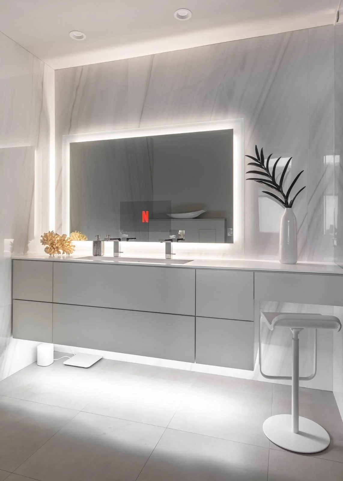 Modern bathroom with white vanity, large mirror with backlighting, decorative vase with black leaves, and minimalistic decor.