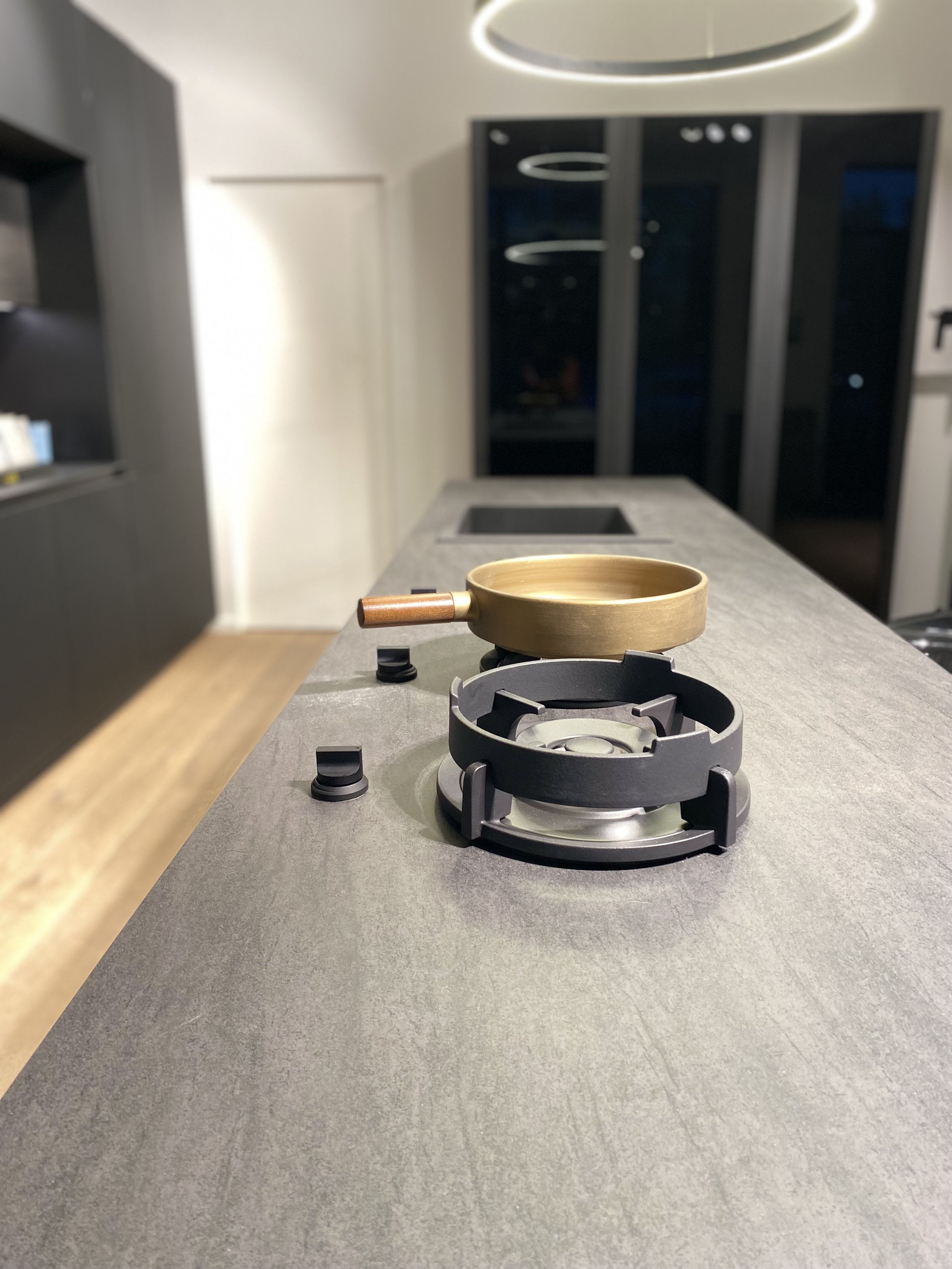 A modern kitchen with a countertop stovetop, featuring a brass pan and a black burner grate, with a kitchen island and sliding glass doors in the background.