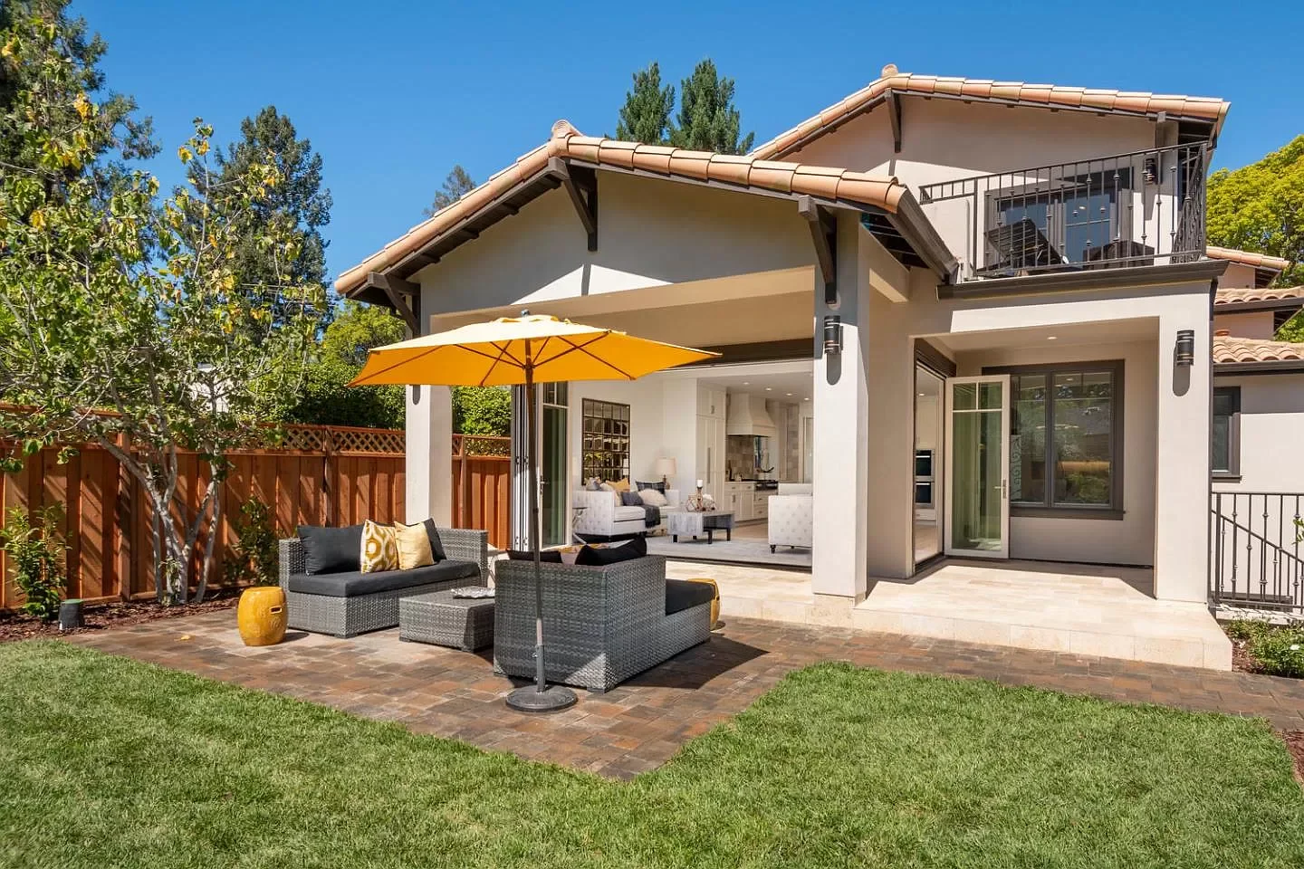 Backyard patio with outdoor seating, umbrella, and views into the house's living room and kitchen, with a yard and fence.