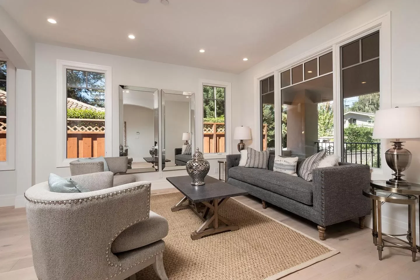 Living room with gray sofa, beige armchair, large windows, mirror, side tables with lamps, decorative vases, and wooden coffee table on a woven rug.