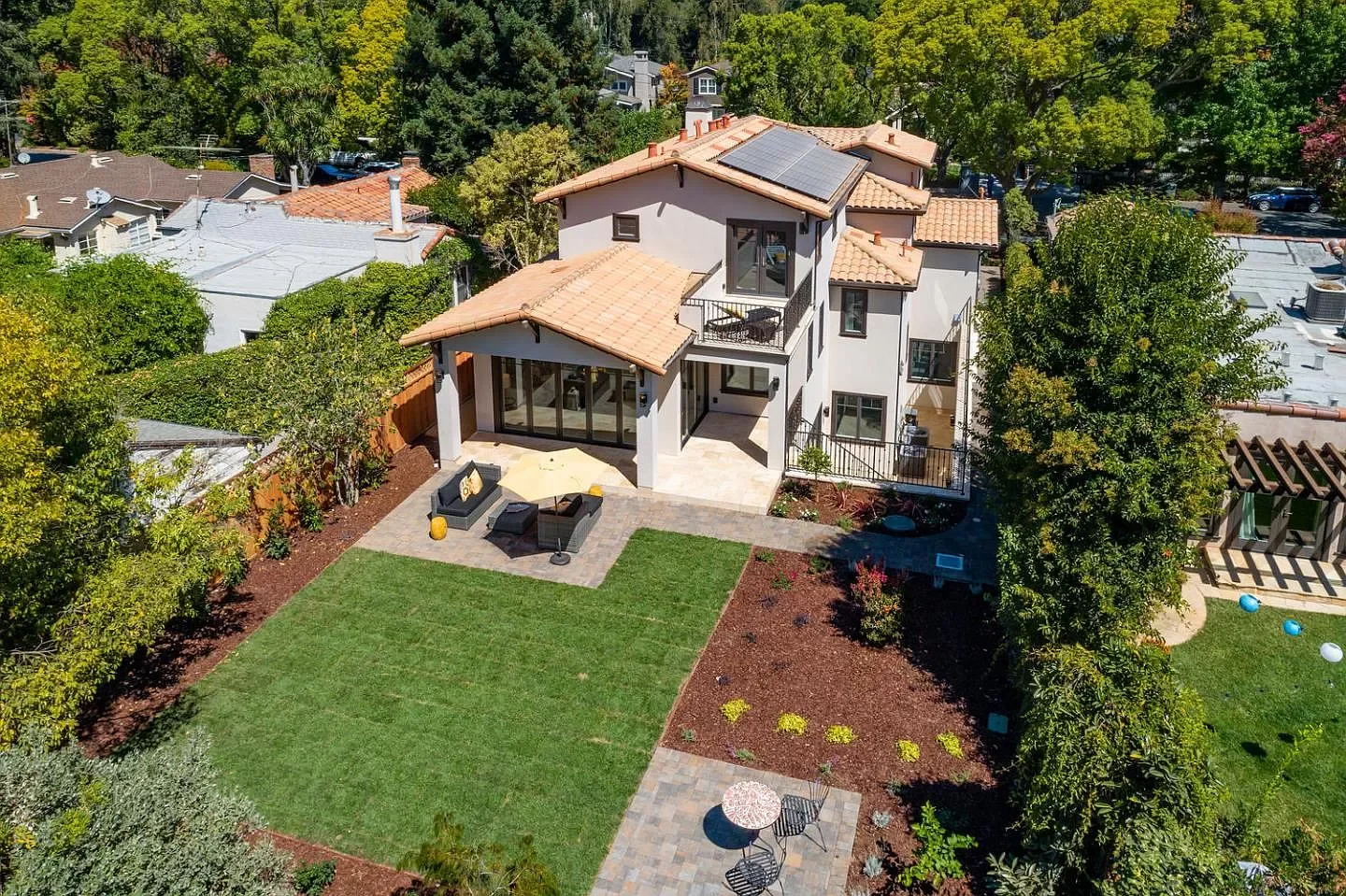 Aerial view of a modern house with solar panels on the roof, surrounded by trees and a landscaped backyard with outdoor furniture, a grassy lawn, and paved patio areas.