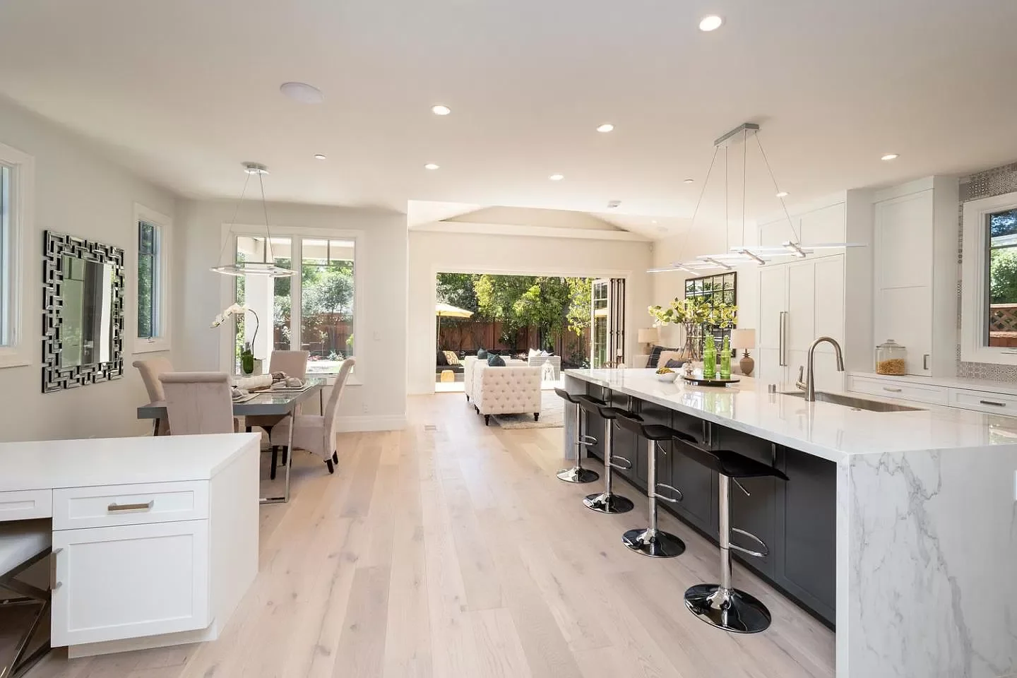 Bright, modern kitchen and dining area with white cabinets, a marble island with three barstools, and an open outdoor patio visible through sliding glass doors.