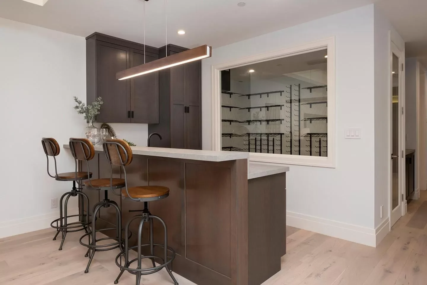 Modern kitchen with dark upper cabinets, white countertops, a bar with three wooden and metal barstools, and a walk-in wine cellar with black racks visible through a large window.