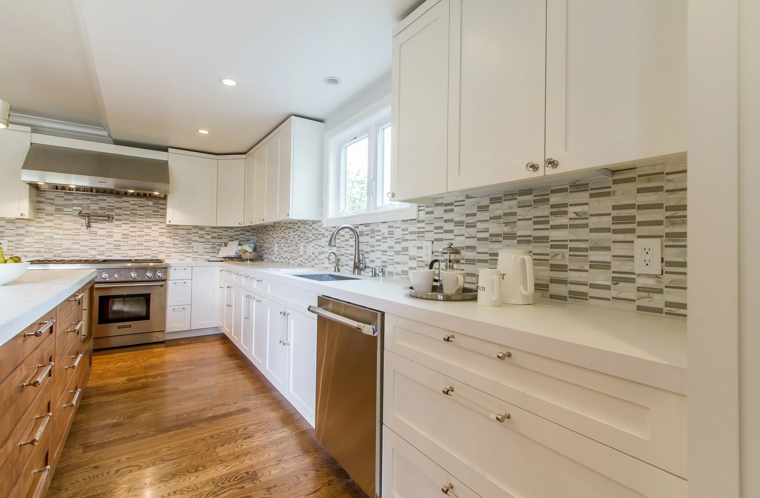 Modern kitchen with white cabinets, a gray tiled backsplash, and stainless steel appliances, including a stove and dishwasher, on warm wooden floors.