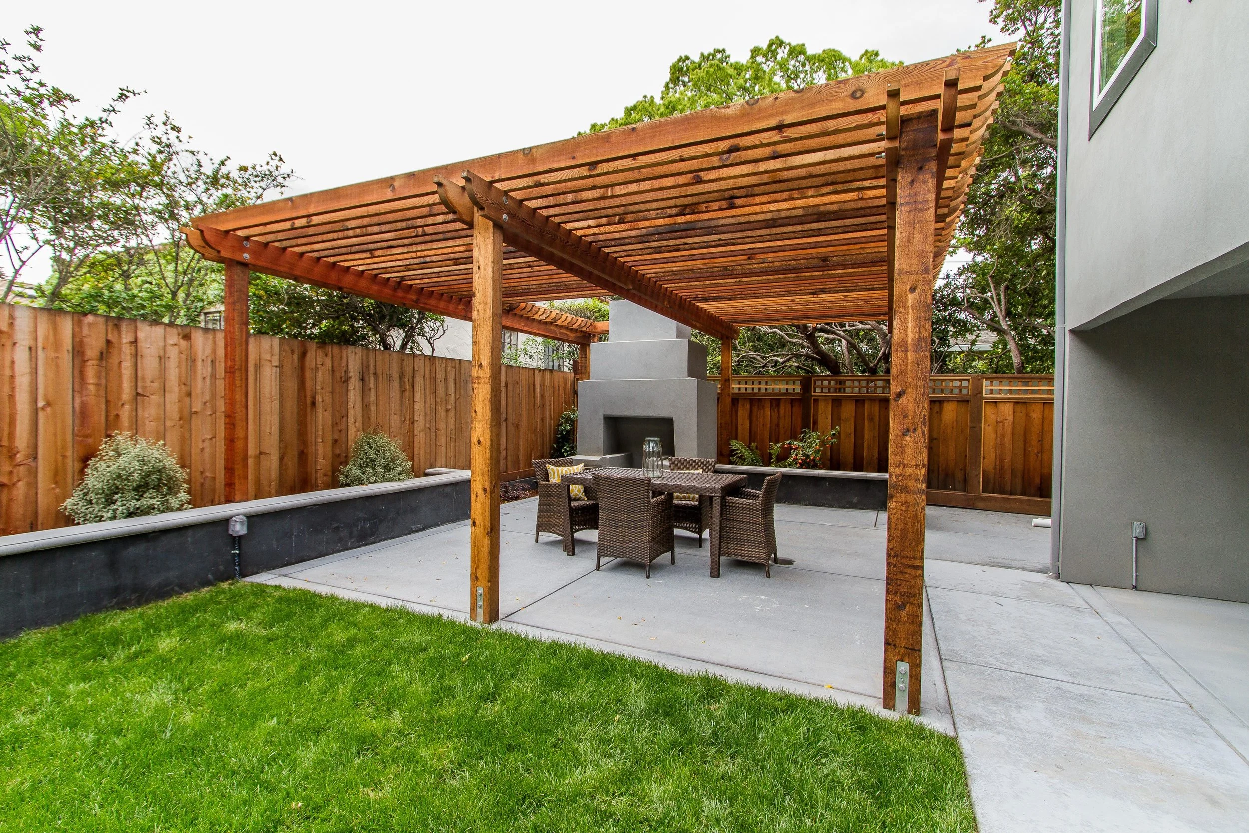 An outdoor patio area with a wooden pergola, a concrete floor, a green lawn, a fenced backyard, outdoor furniture, and a fireplace.