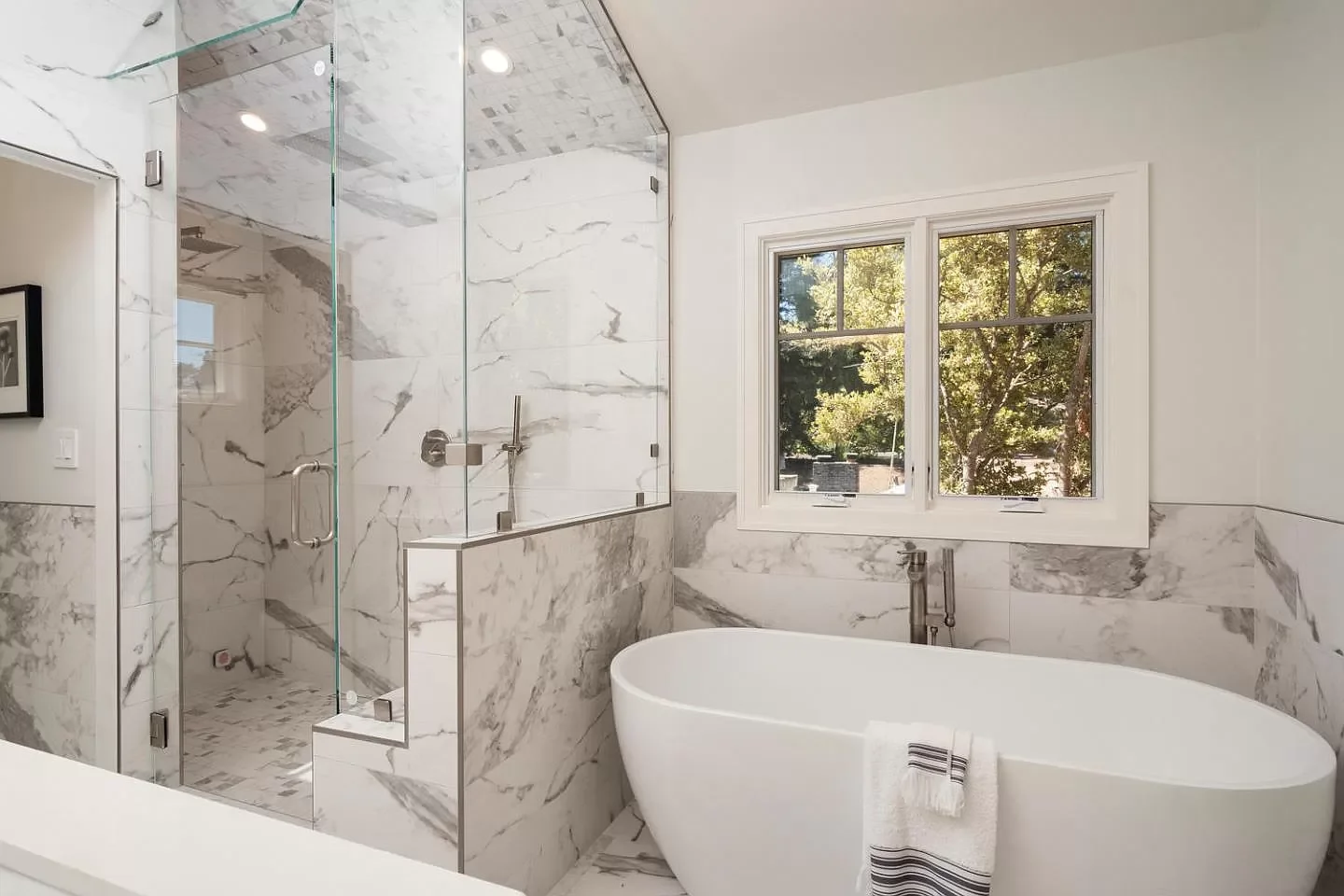 A luxurious bathroom featuring a marble bathtub and a glass-enclosed shower with marble walls, and a window showing trees outside.