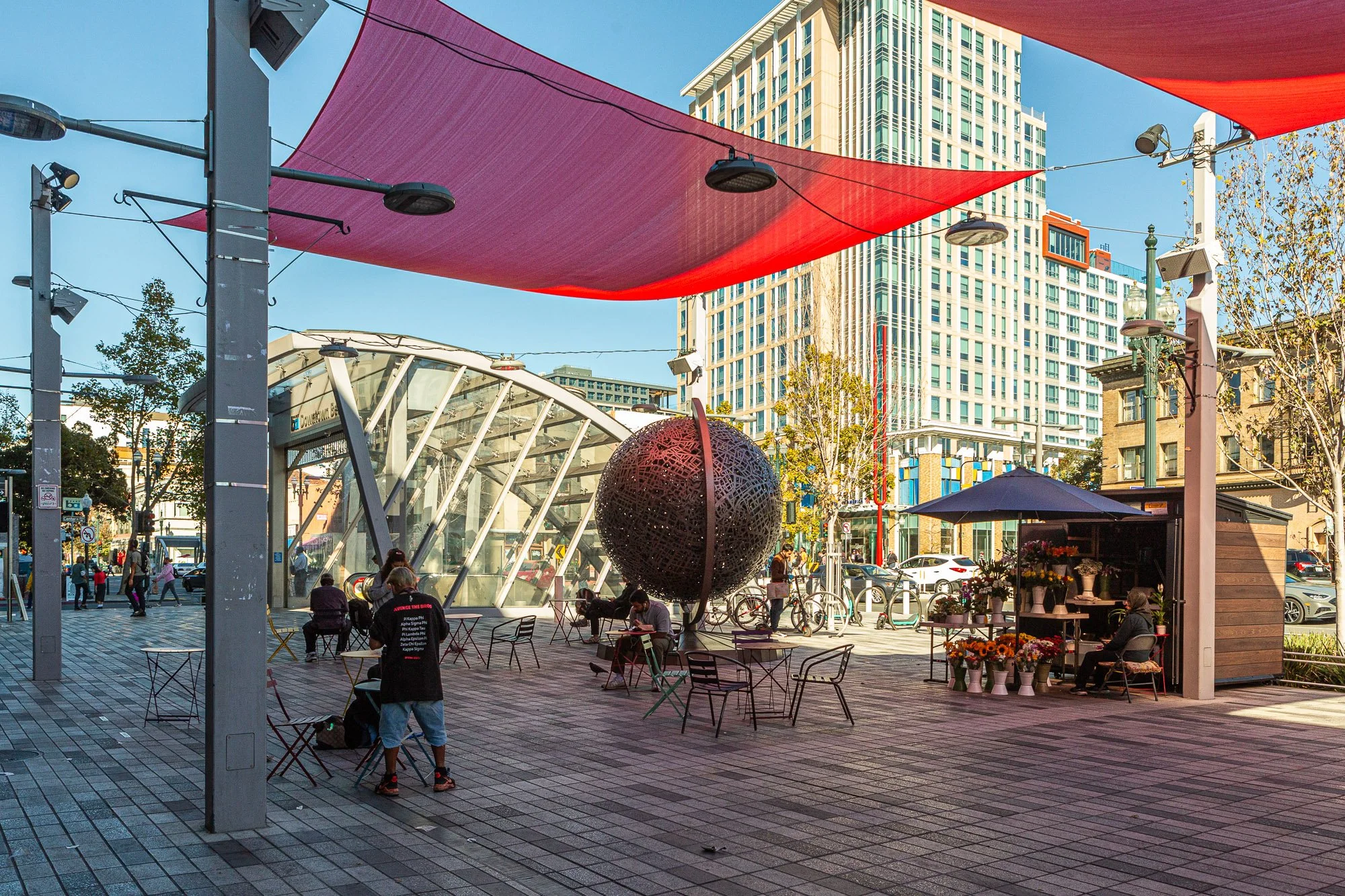 Urban outdoor scene with a modern glass structure, seating area with chairs, people sitting and standing, a large spherical sculpture, storefront with flowers, and high-rise buildings in the background under a blue sky with red fabric shade overhead.