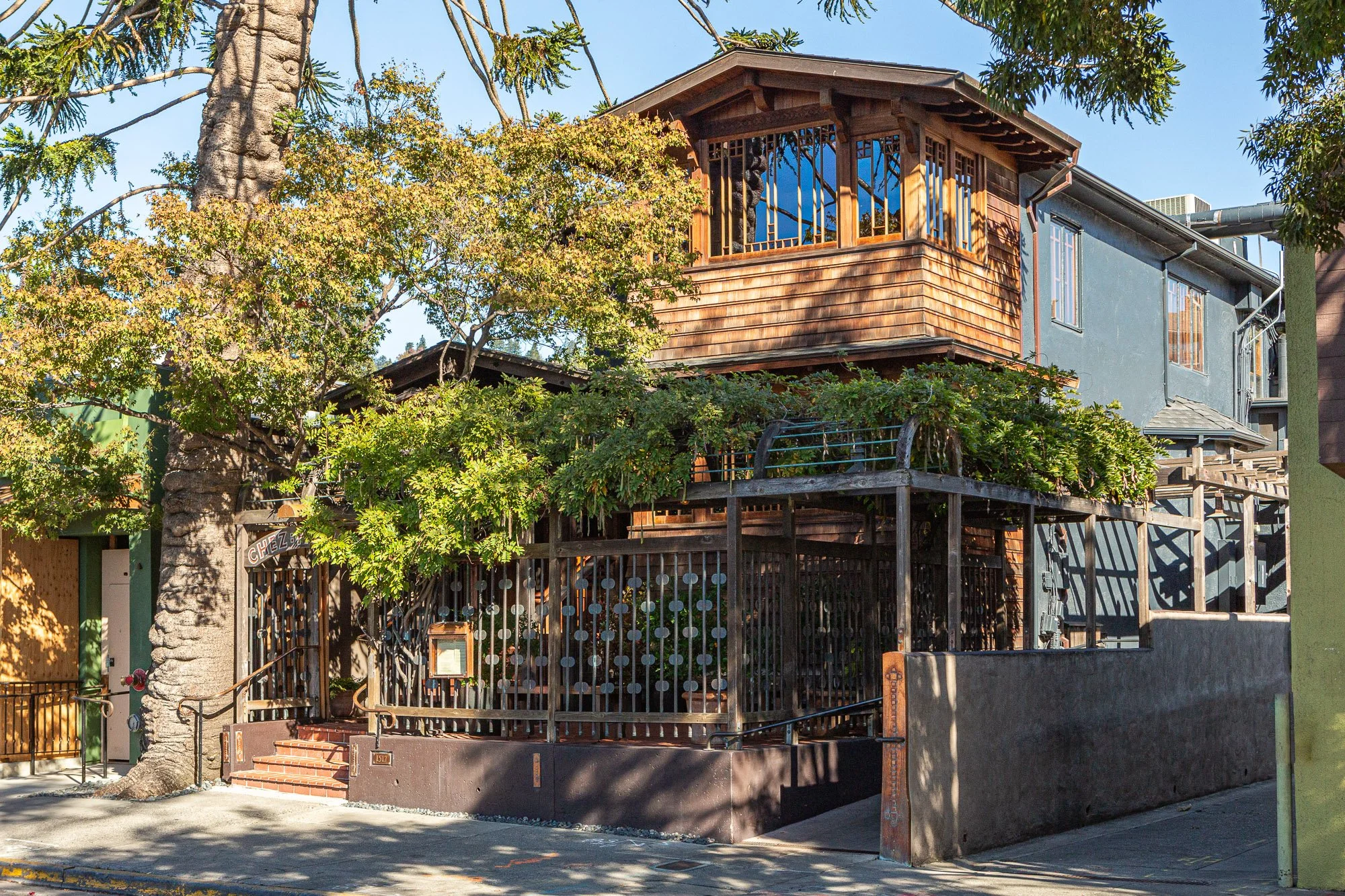 A multi-story house with a wooden upper deck and blue painted lower level, surrounded by trees and greenery, on a city sidewalk.