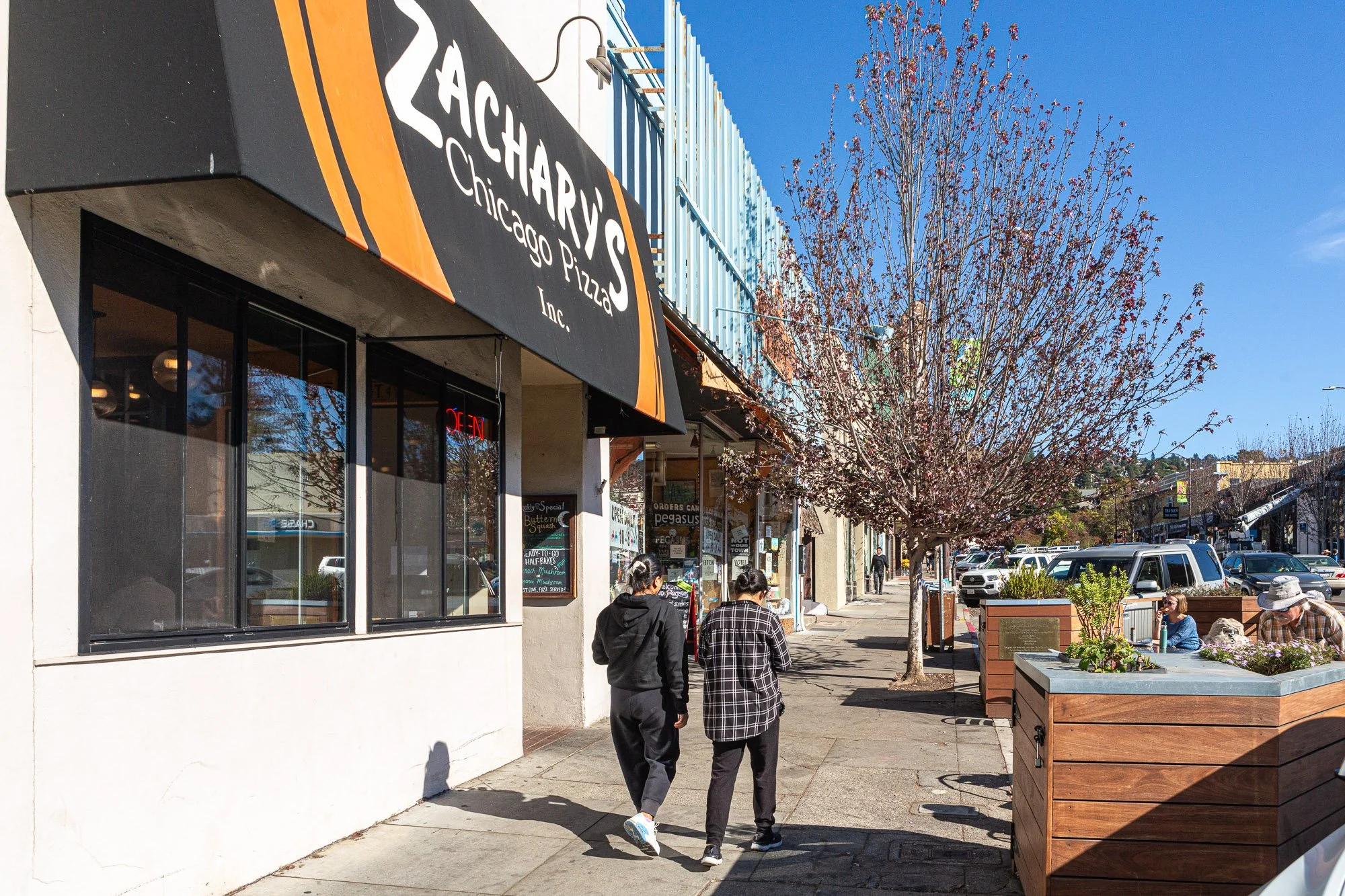People walking on the sidewalk in front of Zachary's Chicago Pizza restaurant on a sunny day, with leafless trees and parked cars visible in the background.