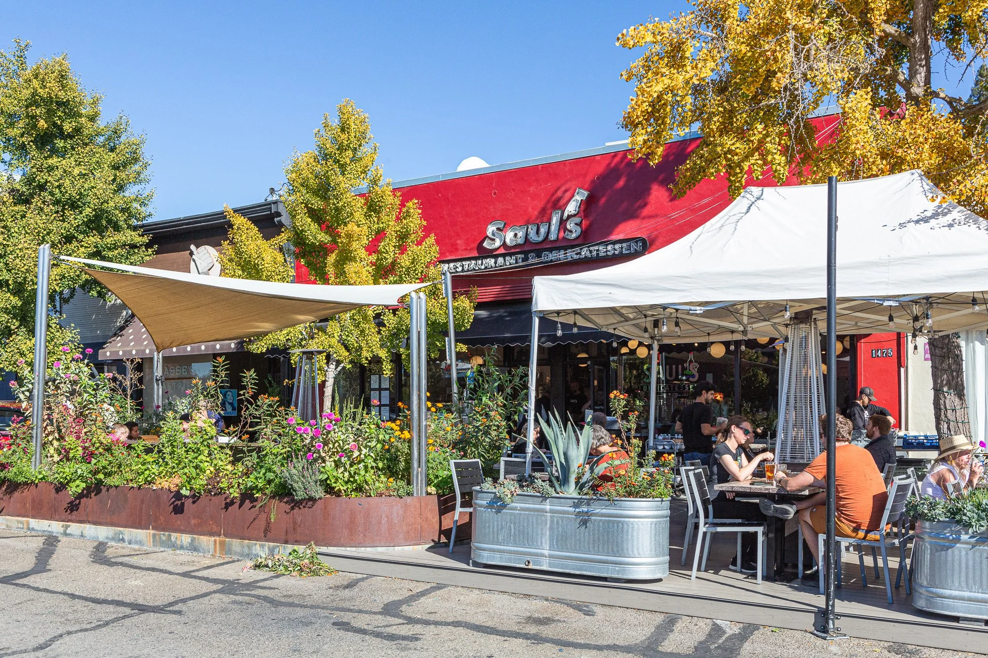 Outdoor patio of Saul's restaurant with people dining, surrounded by potted plants, trees with yellow leaves, and shaded by white and beige canopies on a sunny day.