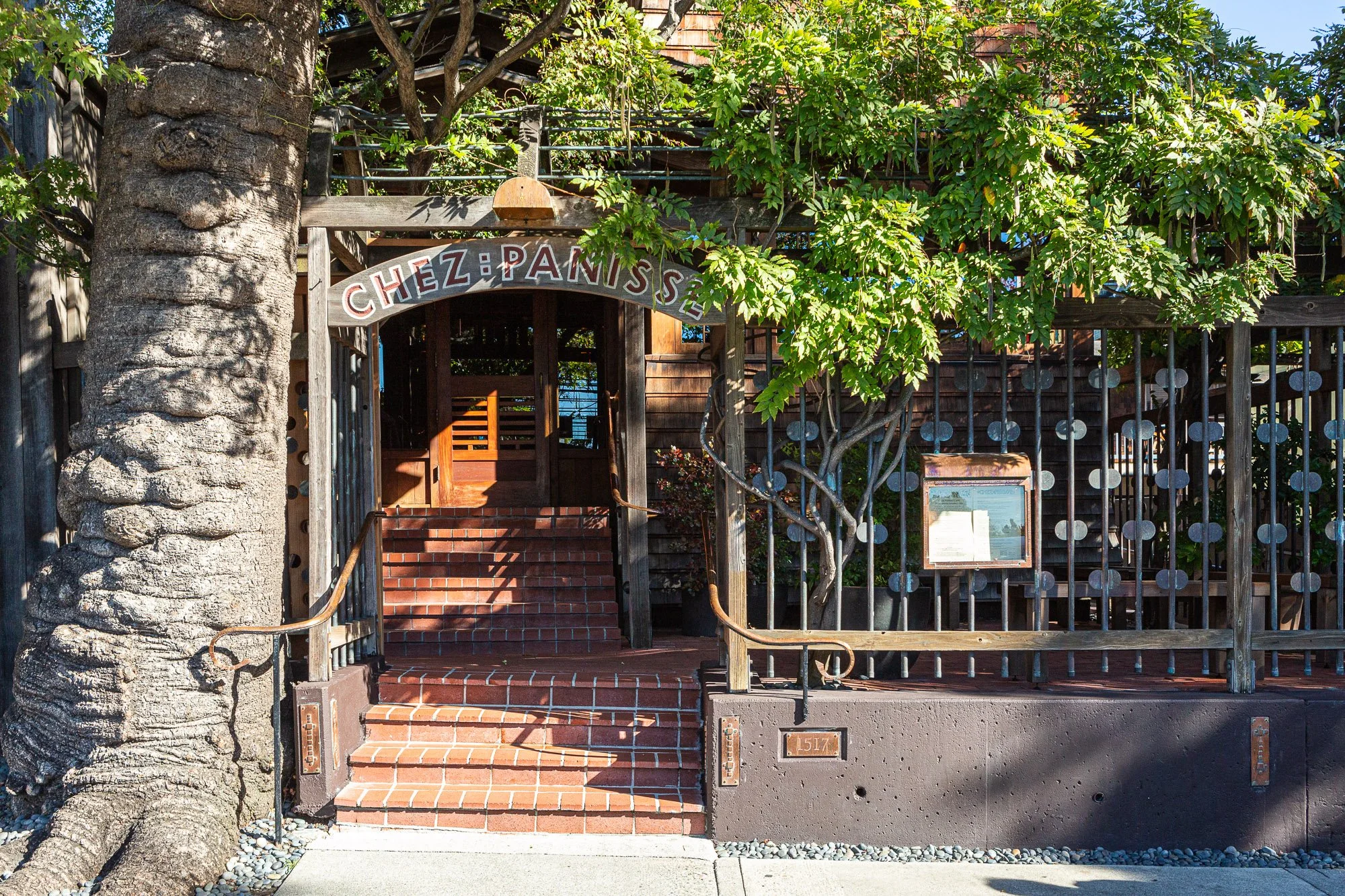 Entrance to Chez Panisse restaurant with brick stairs, wooden archway, trees, and outdoor decor.