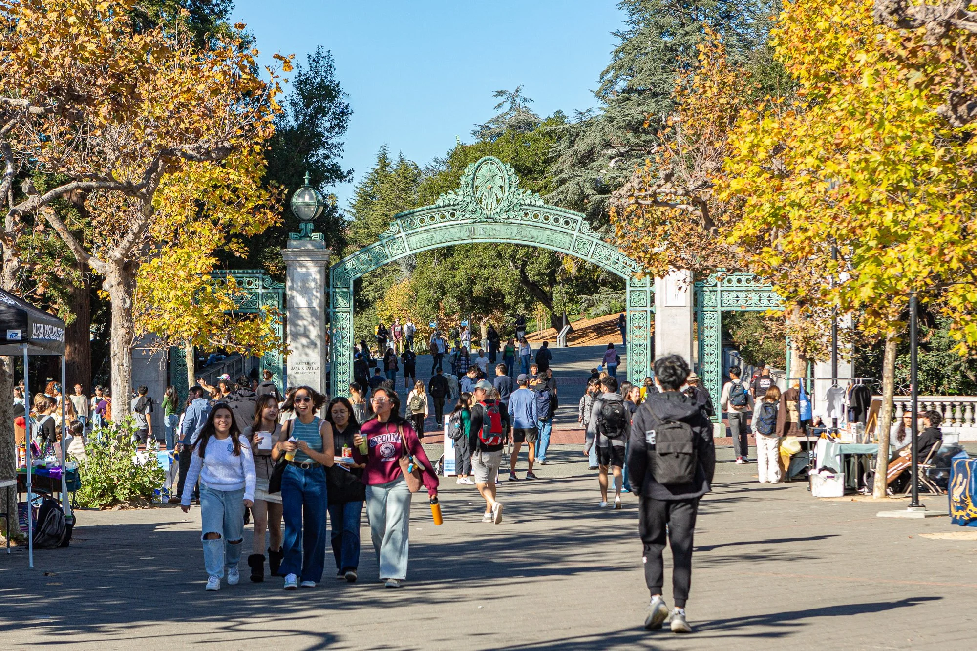 People walking on a pathway under an ornate green metal gate at a university campus, surrounded by trees with autumn foliage.
