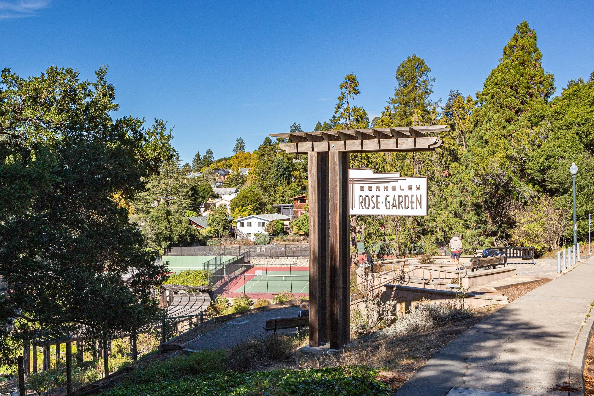 Park with tennis courts, walking path, benches, trees, and a sign reading 'Berkeley Rose Garden' under a clear blue sky.