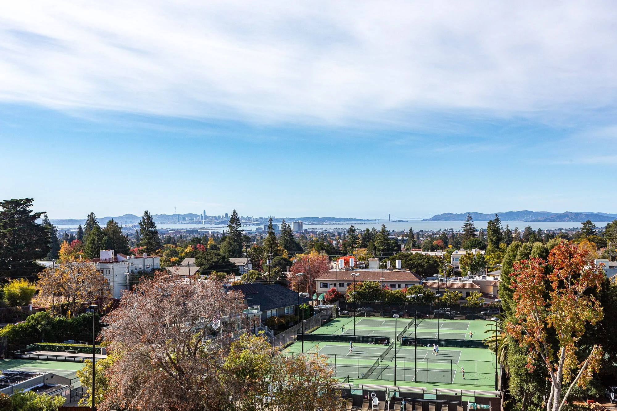 A scenic view of a city skyline, a bay with a bridge, and tennis courts in the foreground with trees and houses surrounding them.